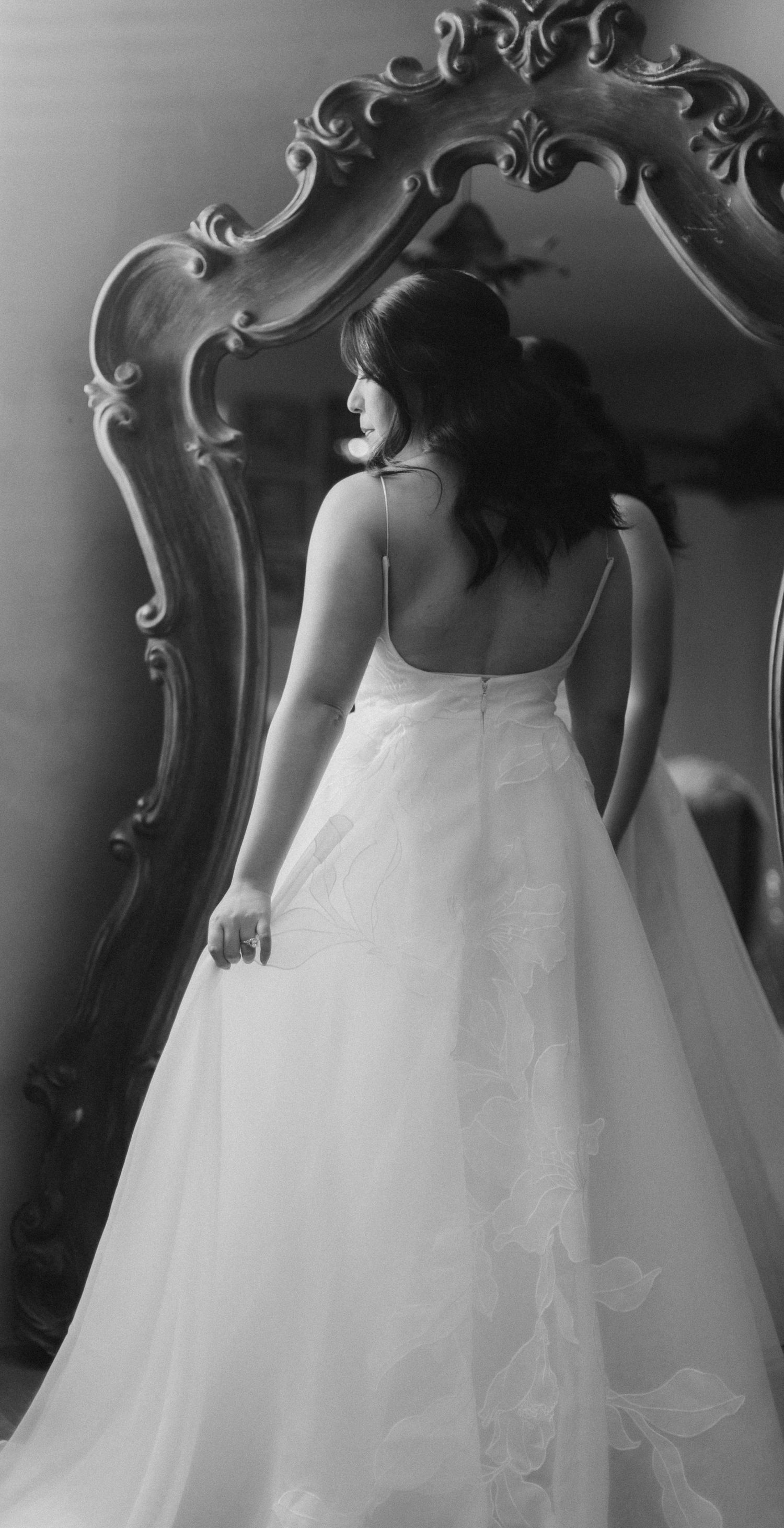 Black and white photo of a bride in a ballgown with a low back standing before an ornate vintage mirror.