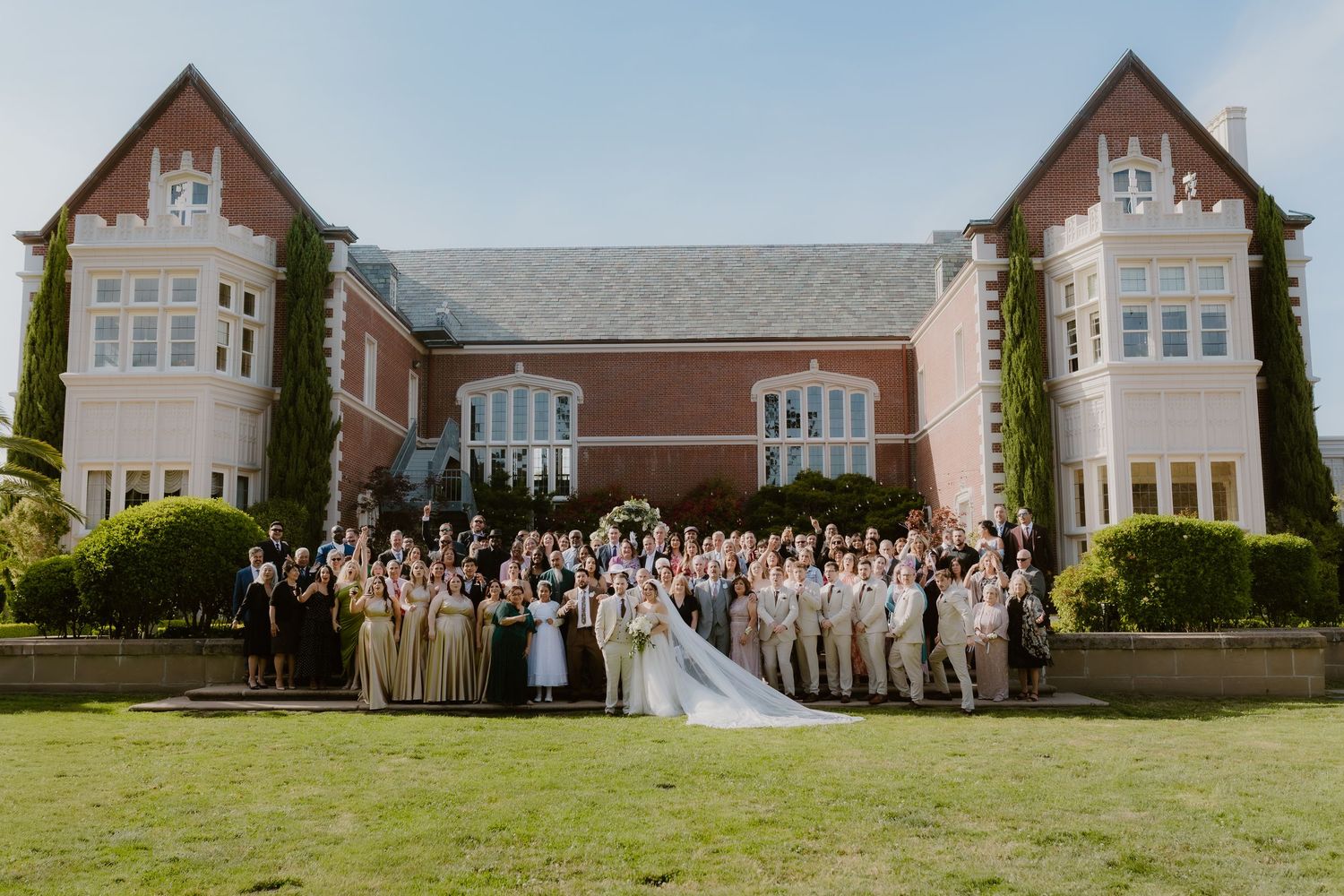 Large wedding party poses in front of elegant brick mansion with ivy-covered walls on sunny day.