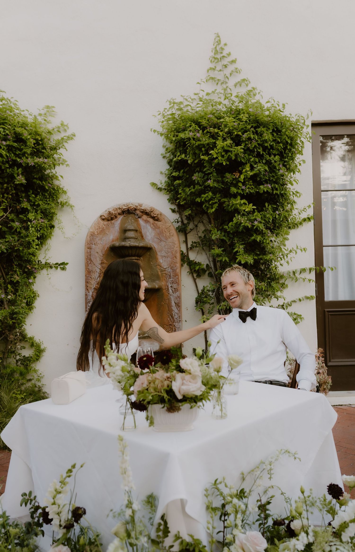 Bride and groom share a joyful moment at their floral-adorned sweetheart table in an elegant outdoor courtyard.