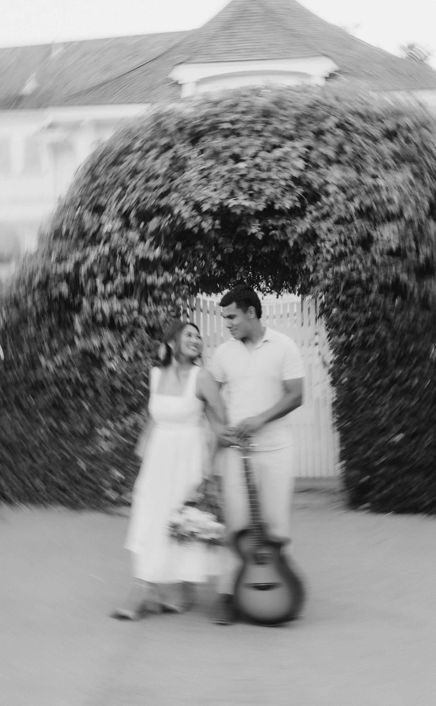Black and white photo of couple standing near ivy arch with guitar leaning against wall.