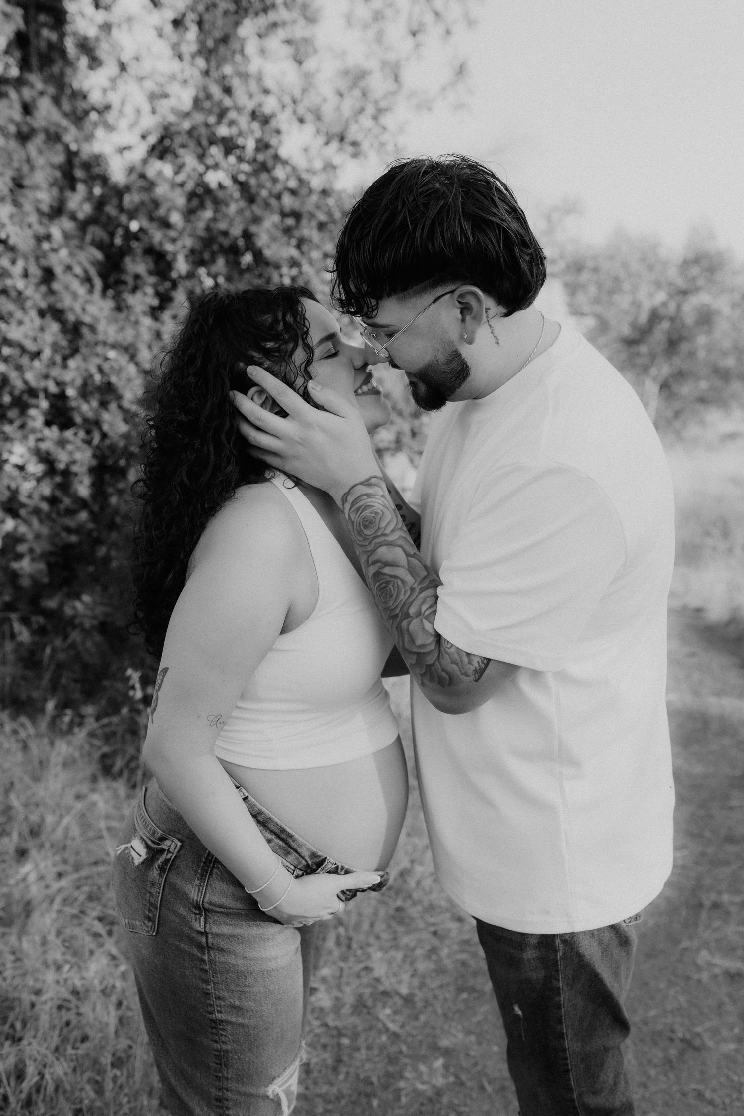 Black and white photo of a tattooed couple sharing a kiss while cradling a pregnant belly outdoors.