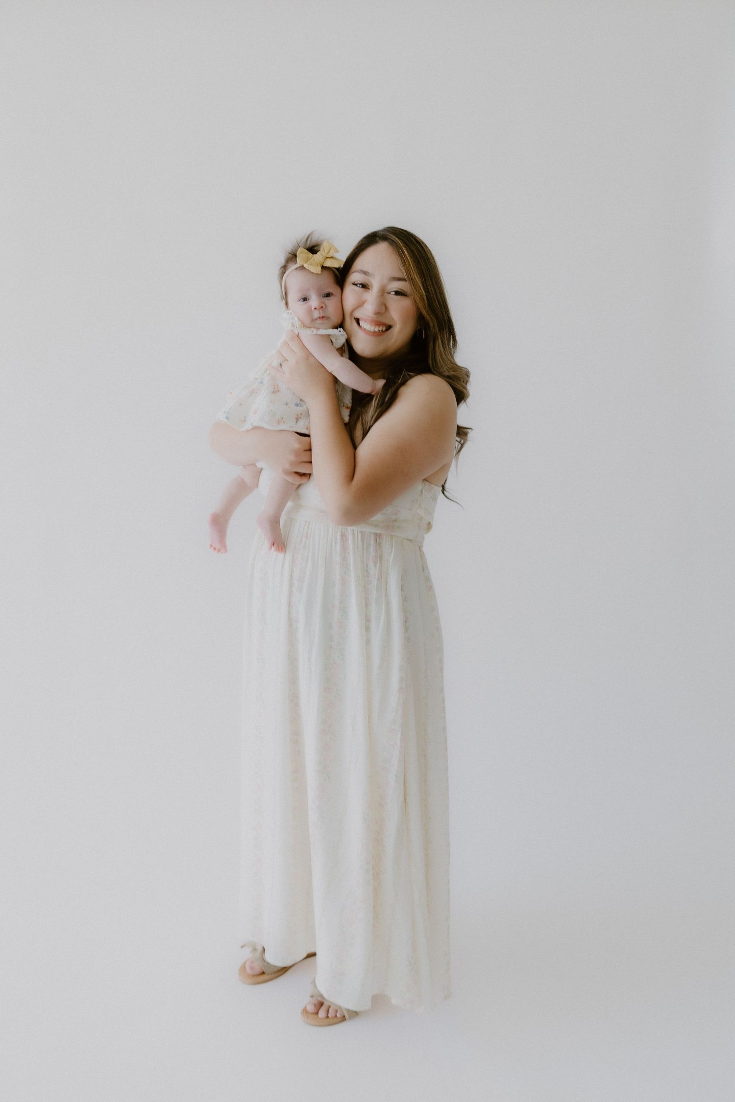 Full-length color photo of a young woman in a white dress holding a baby girl with a yellow bow in a bright studio.