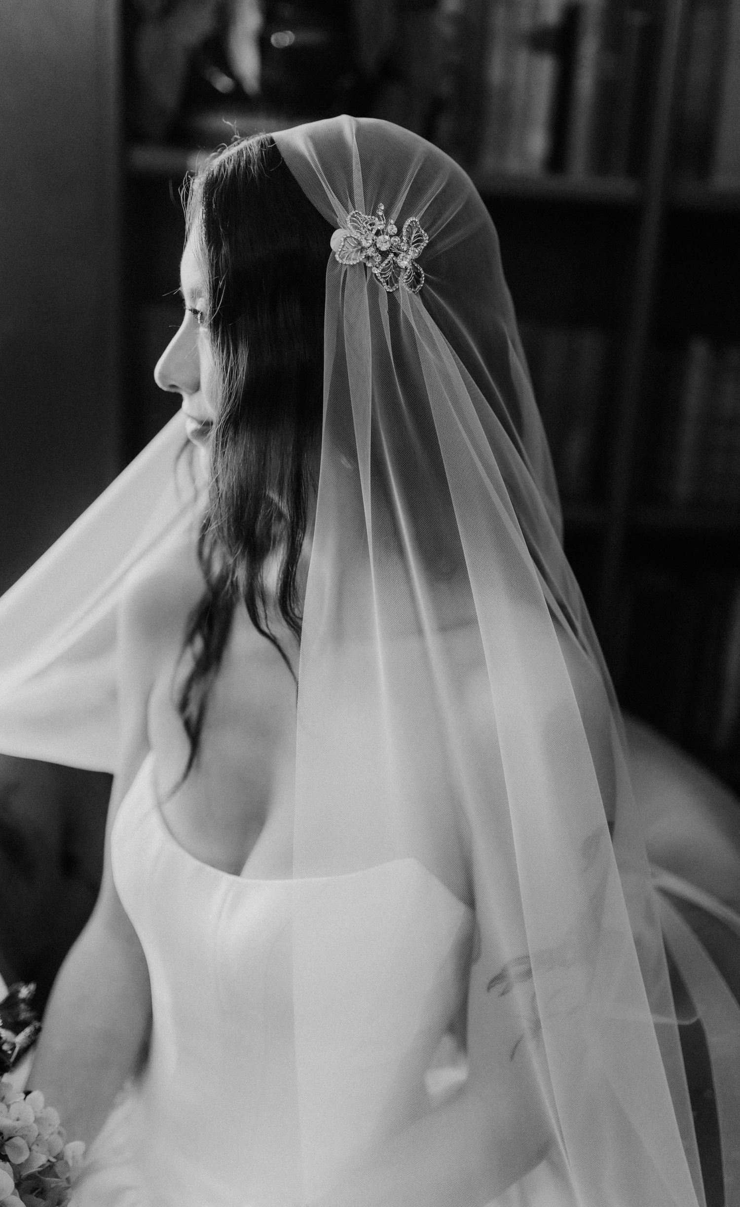 Black and white close-up of bride wearing floral hair comb beneath sheer wedding veil in library.