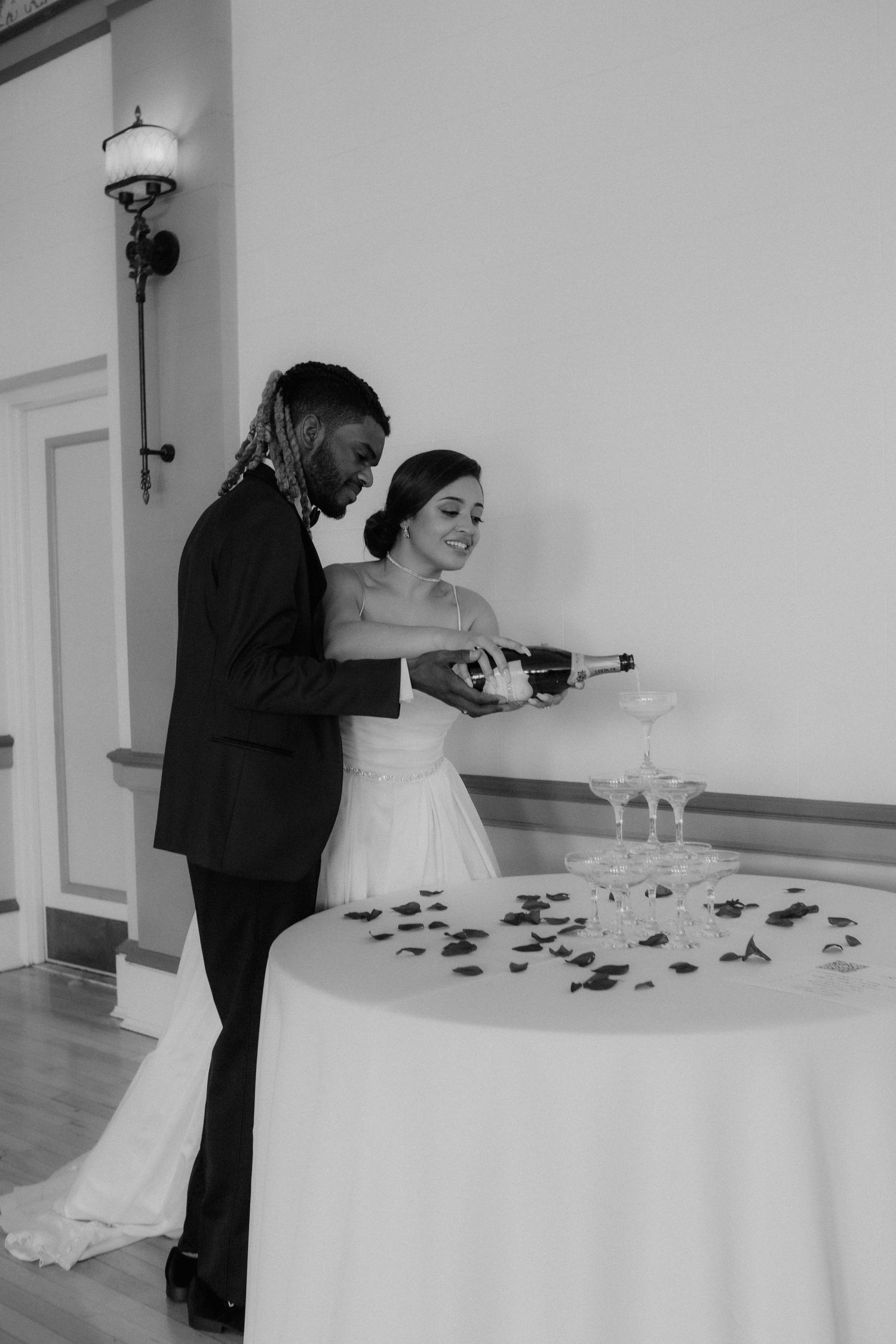 Black and white photo of newlywed couple pouring champagne tower together at elegant wedding reception.