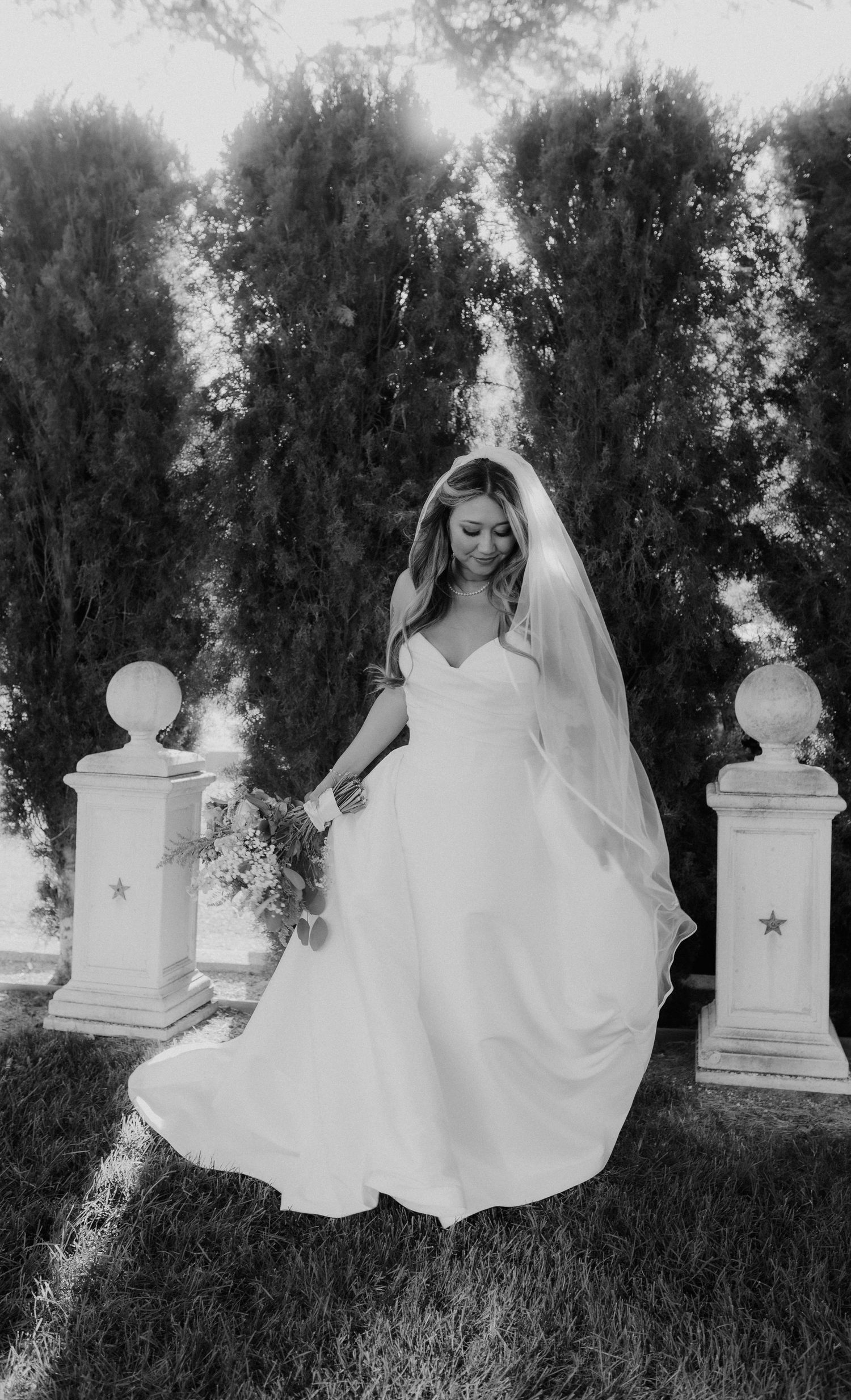 Black and white photo of bride in flowing gown and veil twirling among garden pillars.