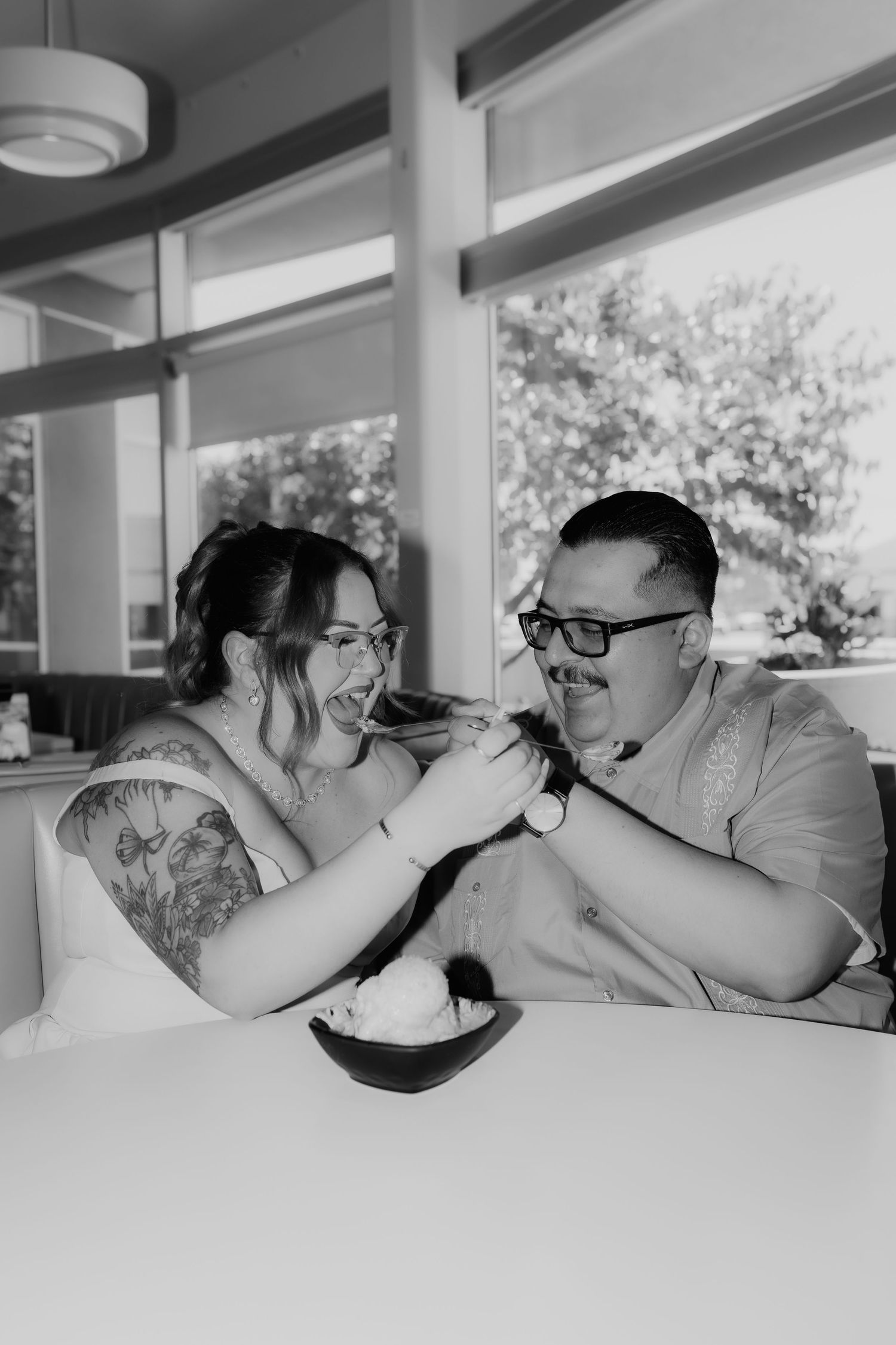 Black and white photo of couple feeding each other ice cream and laughing at retro diner booth.