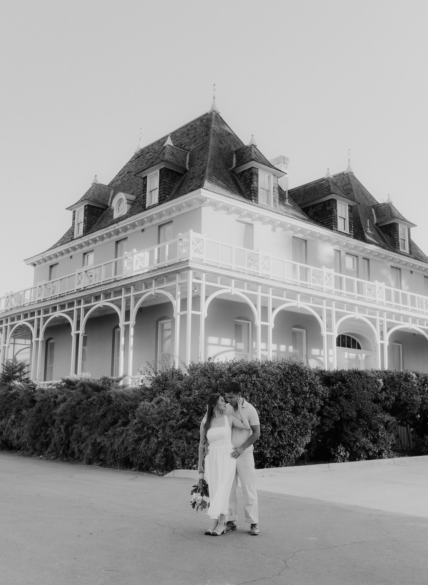 Black and white photo of an engaged couple kissing in front of a grand Victorian mansion exterior.