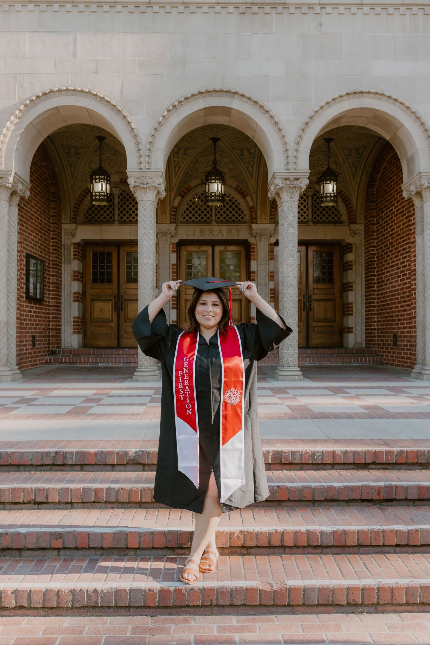 Graduate in black cap and gown with red sash poses on steps of brick building with arched entrance. Fresno Pacific University, California
