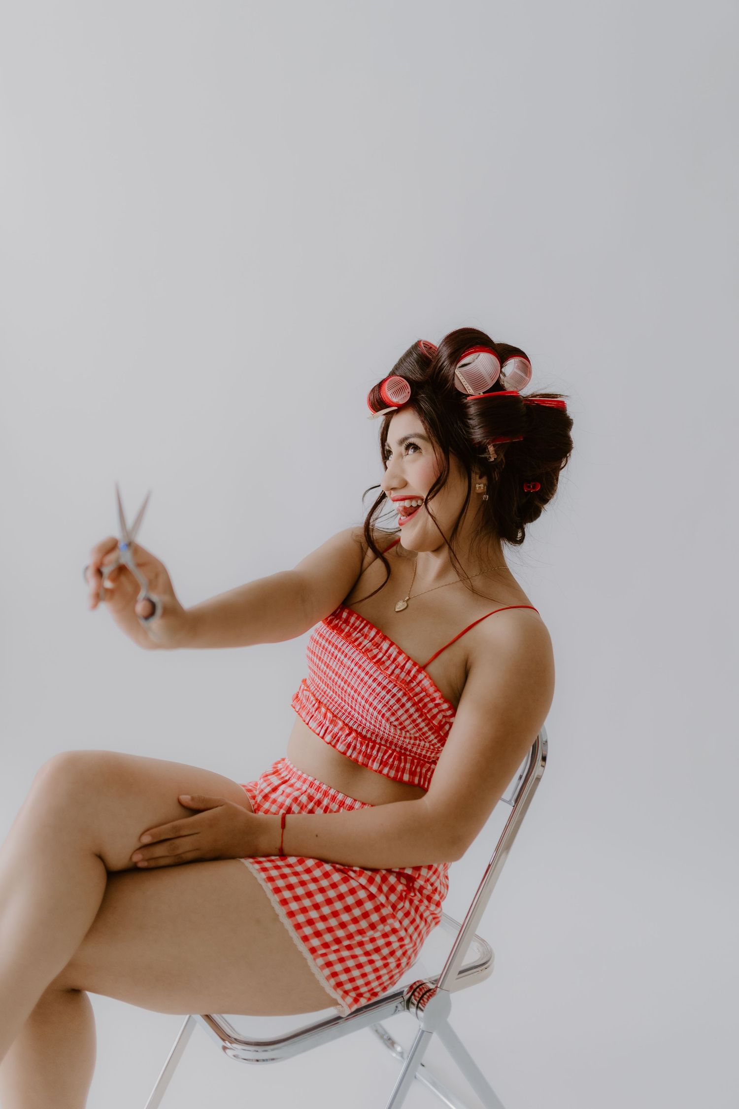 Woman in red gingham two-piece set with hair rollers holds scissors while seated on chrome chair. Branding, Tulare, California