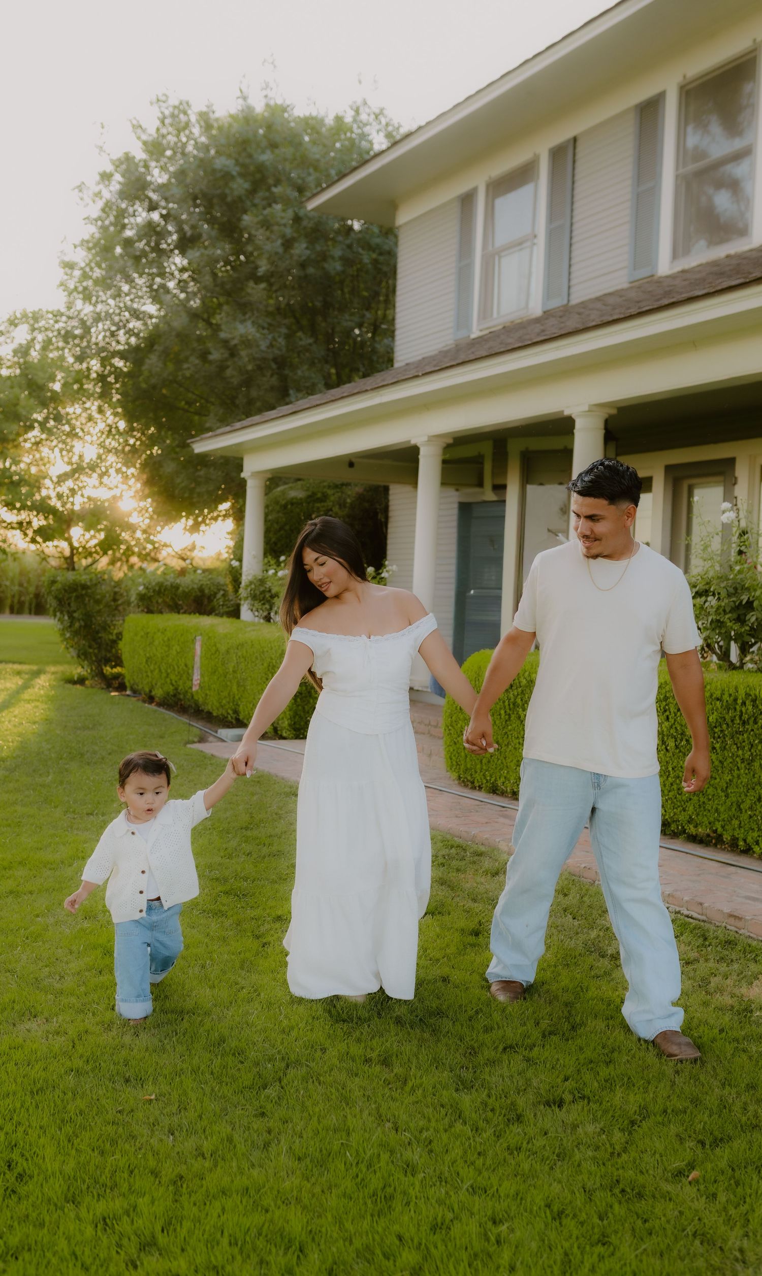 A family of three holds hands walking on green grass in front of a white house at golden hour California charm house Hanford