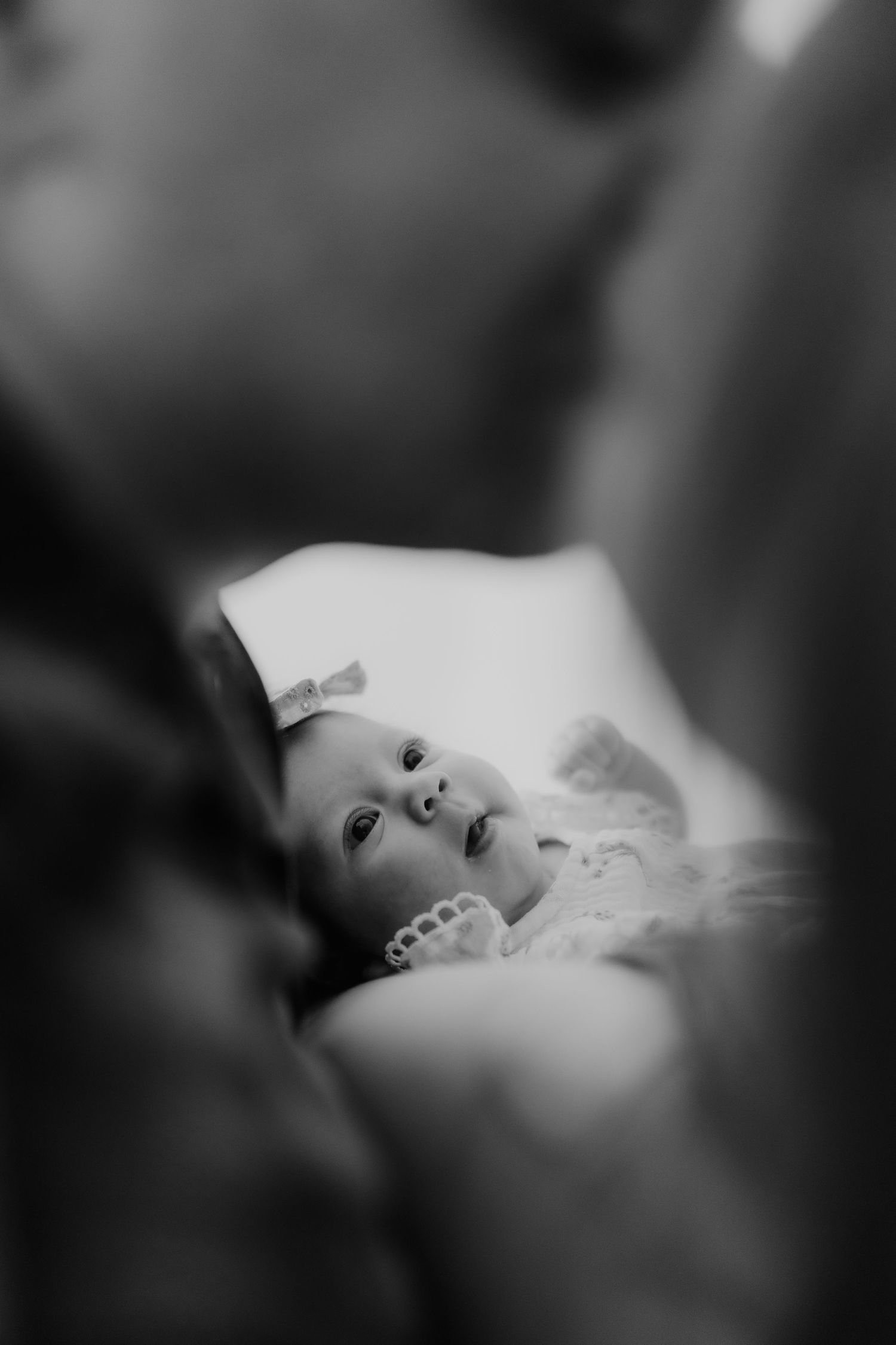Black and white close-up of a newborn baby girl with bow gazing up between two blurred adults.