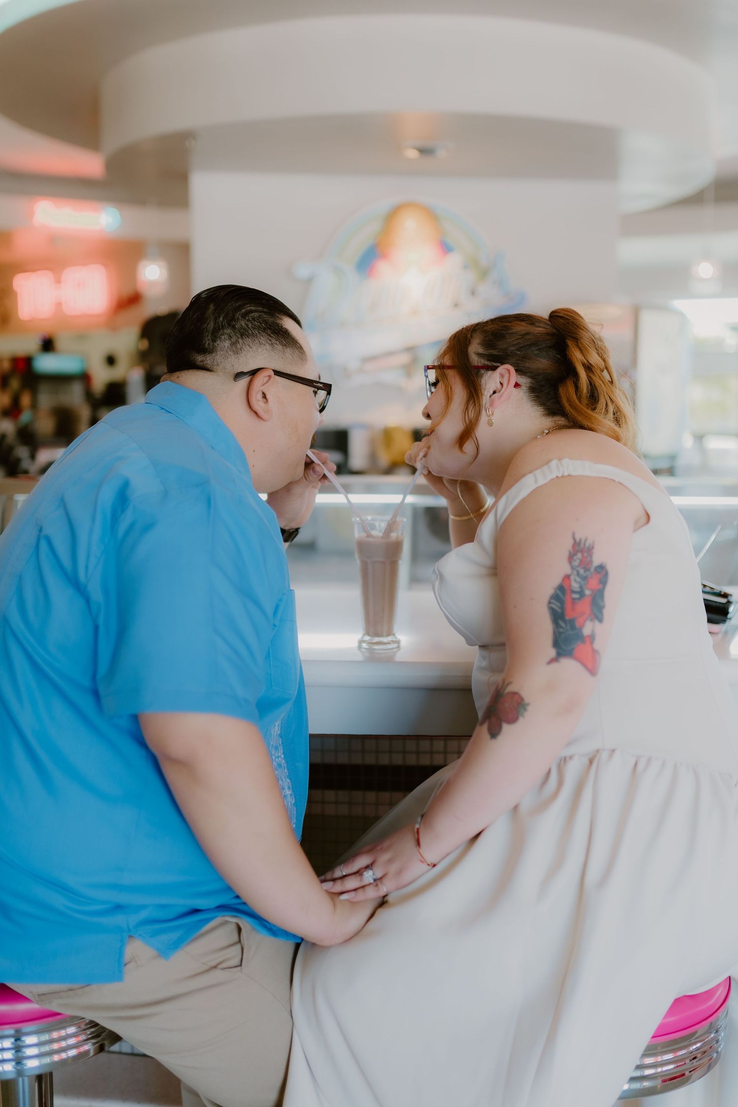 Couple sharing an intimate moment at a retro diner counter, woman in white dress with tattoos, man in blue shirt.