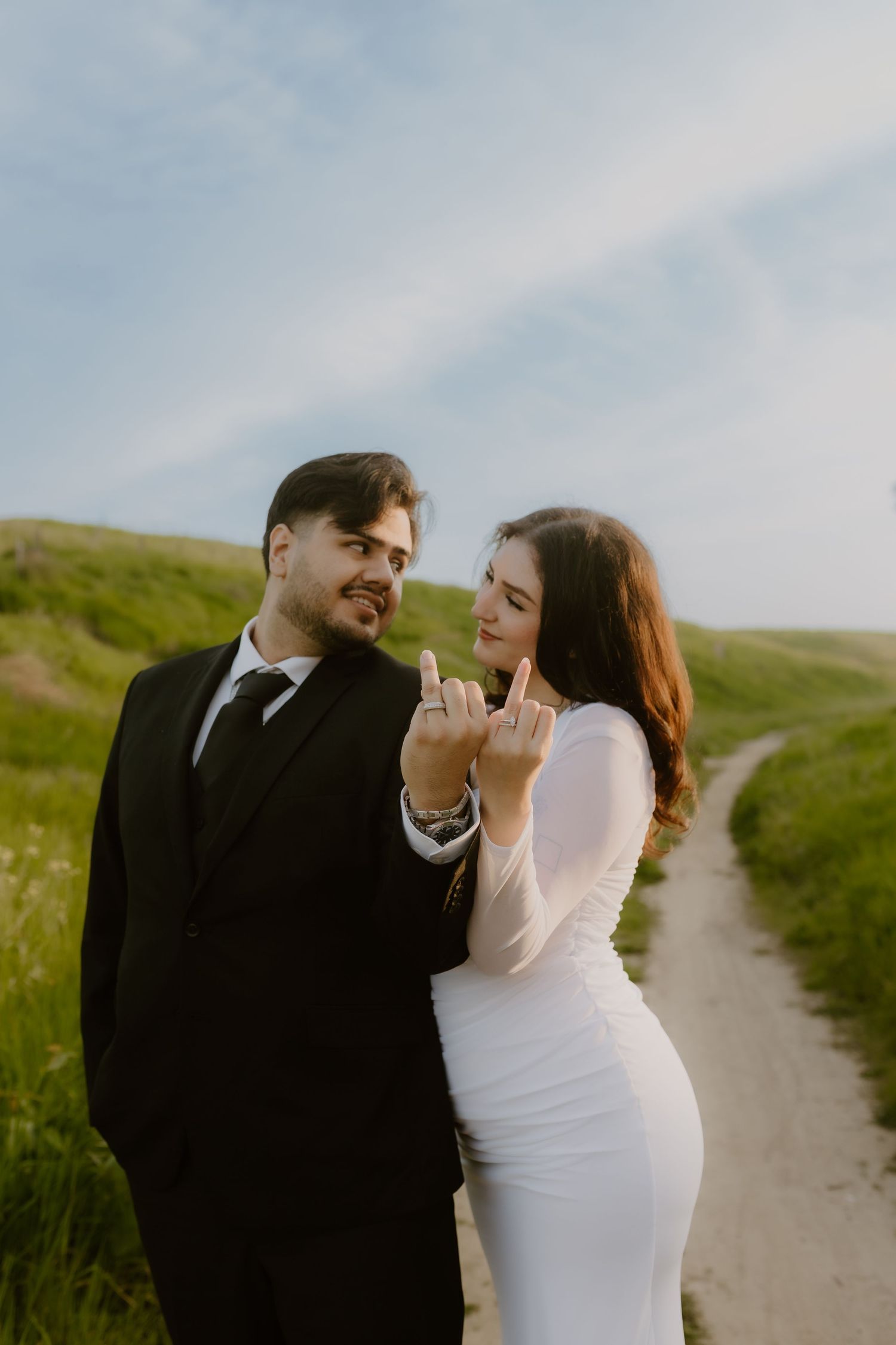 Couple in wedding attire on grassy path showing off wedding rings while smiling at each other.
