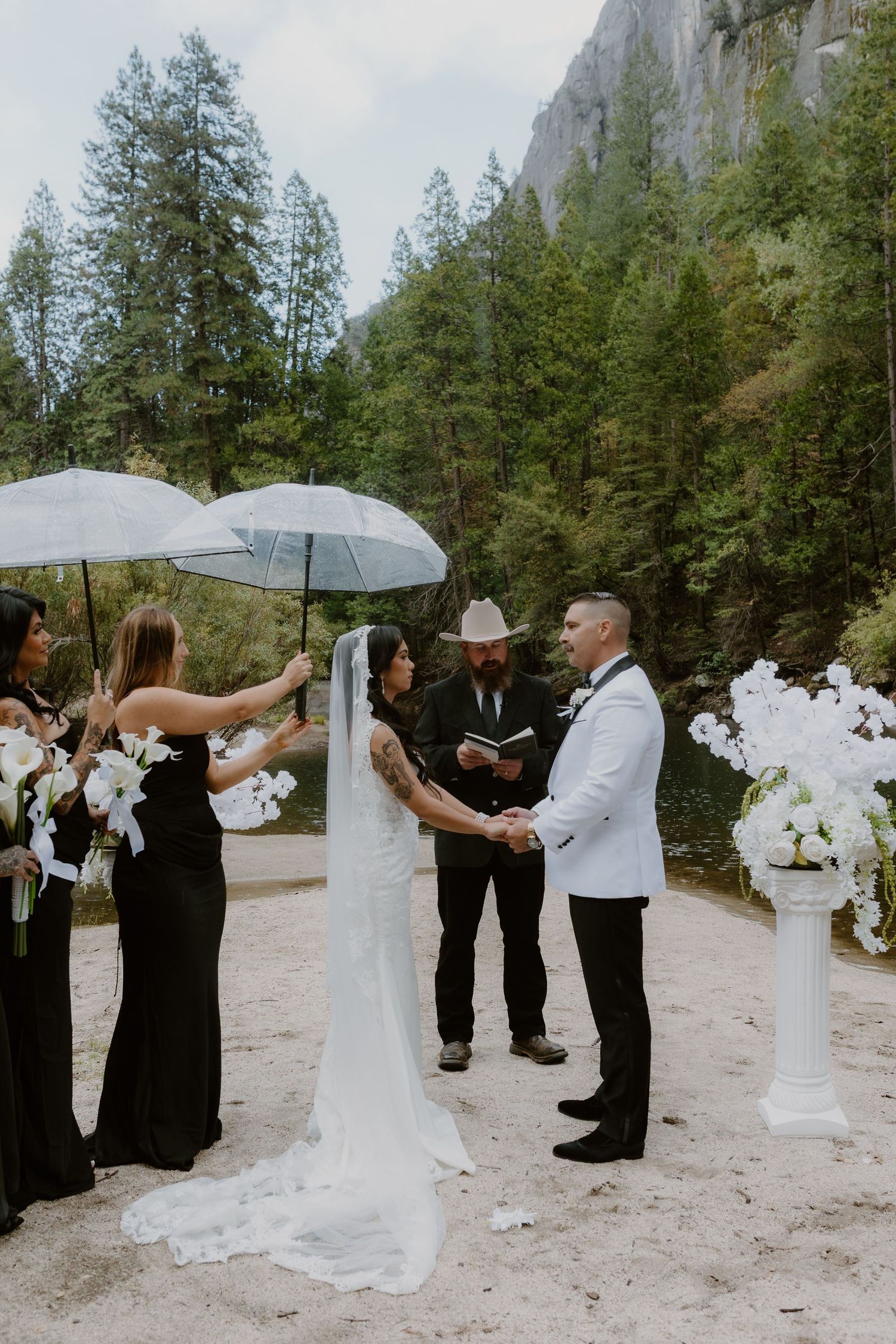 Bride and groom exchanging vows at outdoor Yosemite wedding ceremony with white floral arrangements and clear umbrellas.
