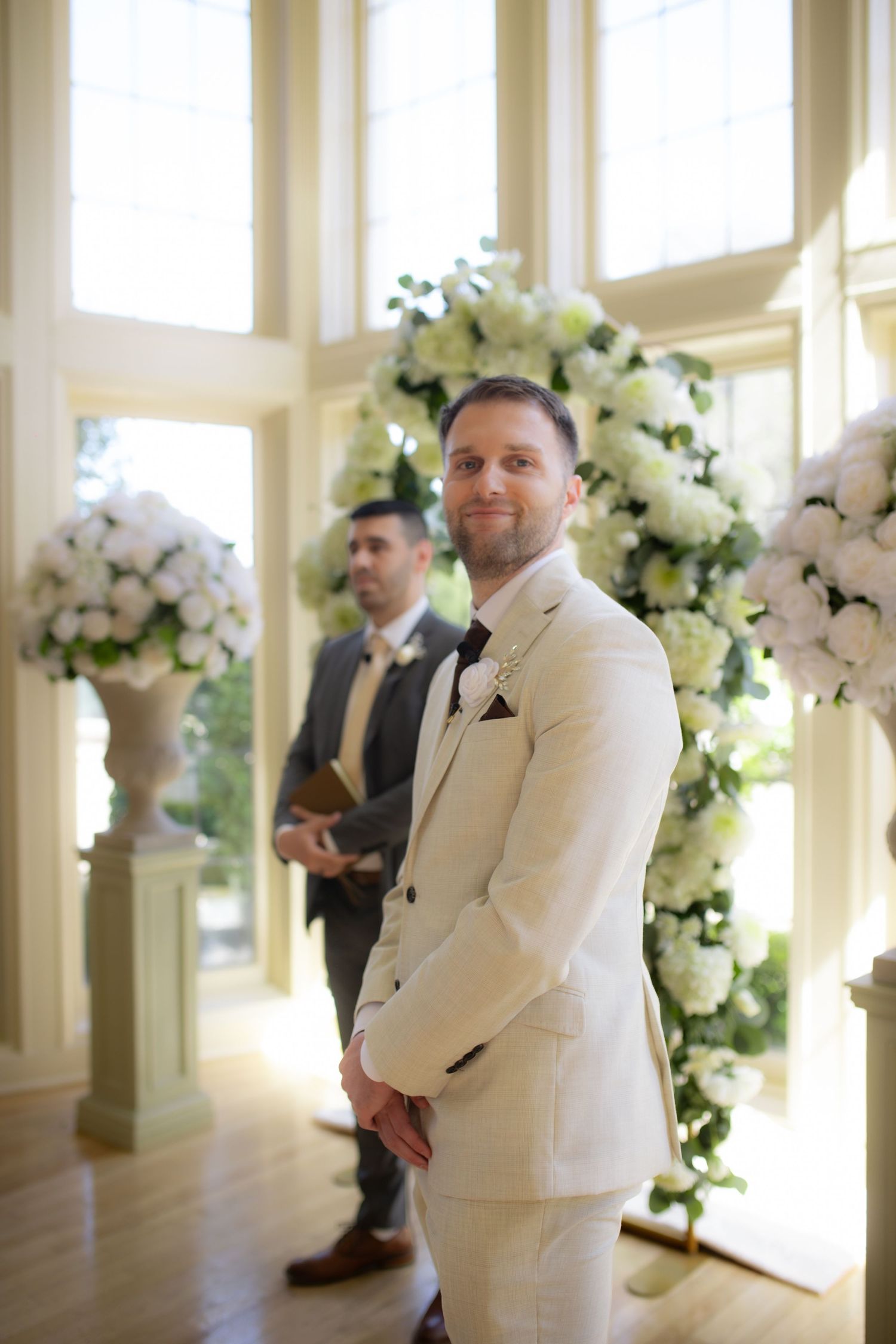 Handsome groom in ivory suit stands at floral altar awaiting bride during elegant wedding ceremony.