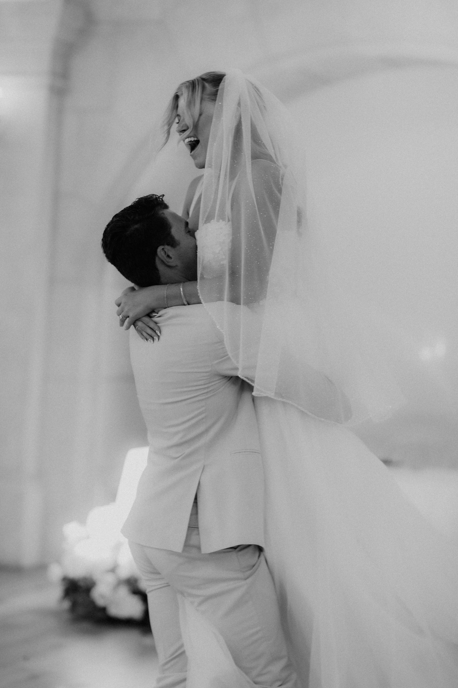 Black and white photo of groom lifting laughing bride in a joyful wedding embrace.