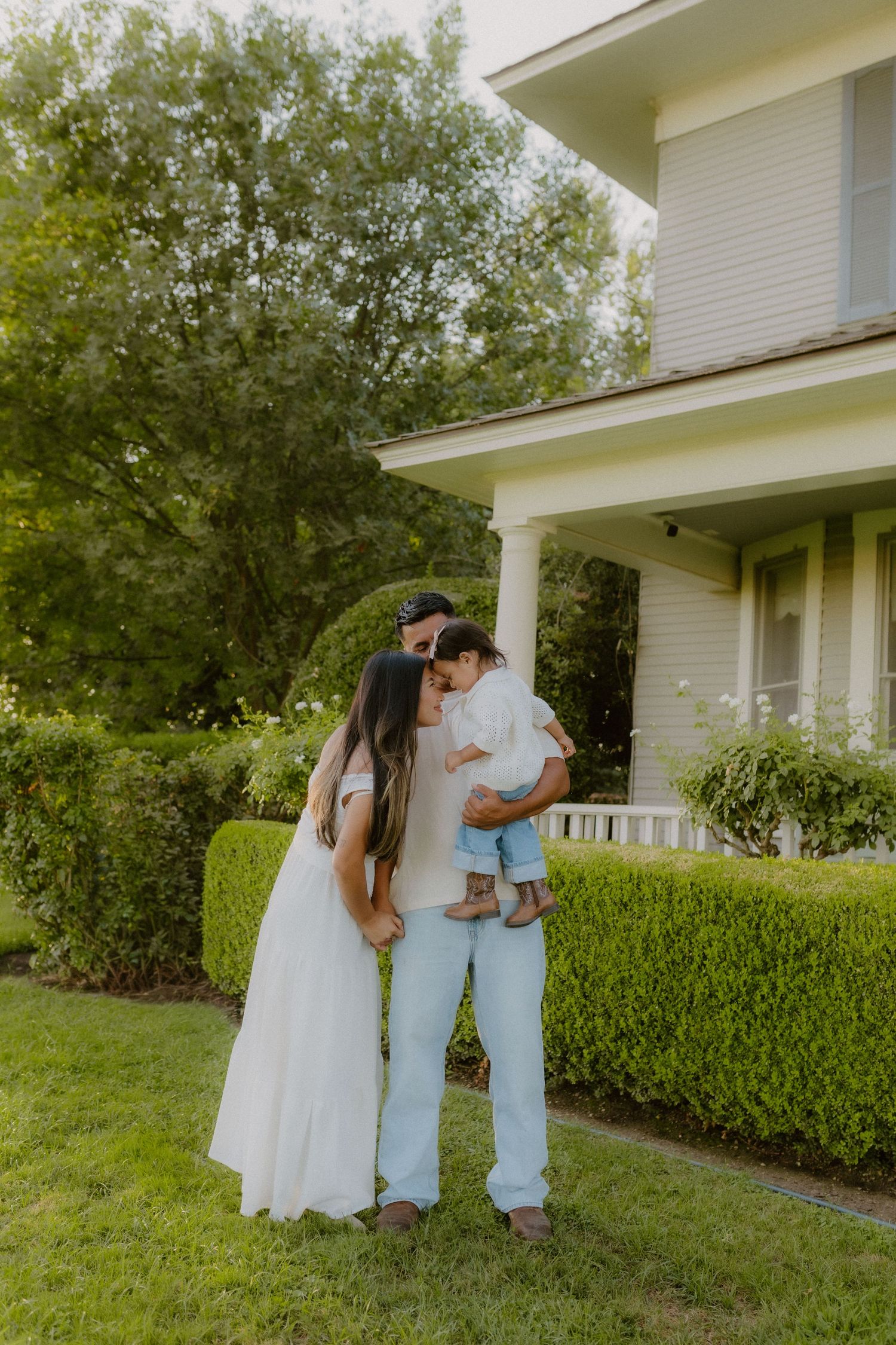A family in white outfits shares a tender moment outside a charming white house surrounded by lush green hedges. Hanford California
