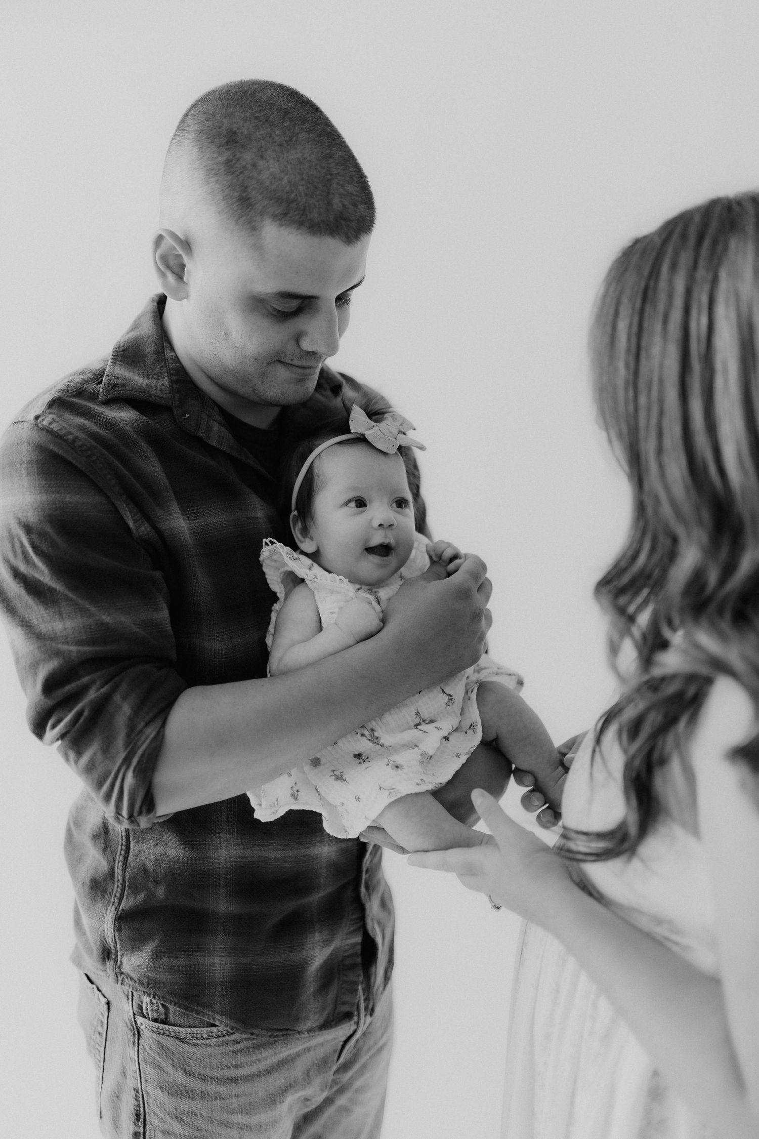 Black and white photo of a father in a plaid shirt holding a baby girl while the mother reaches out to touch her.