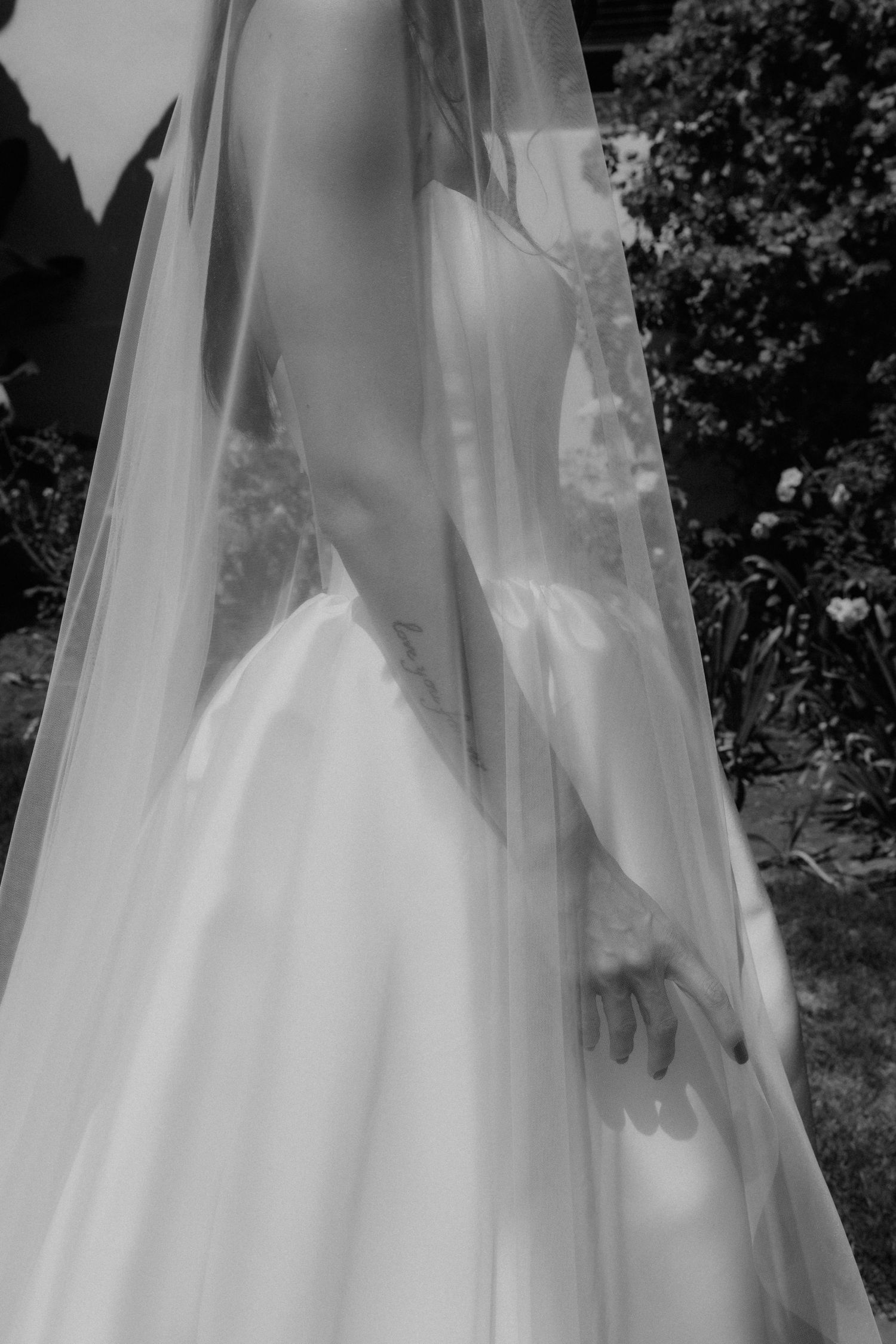 Black and white close-up of bride in flowing wedding gown and veil standing in sunlit garden.