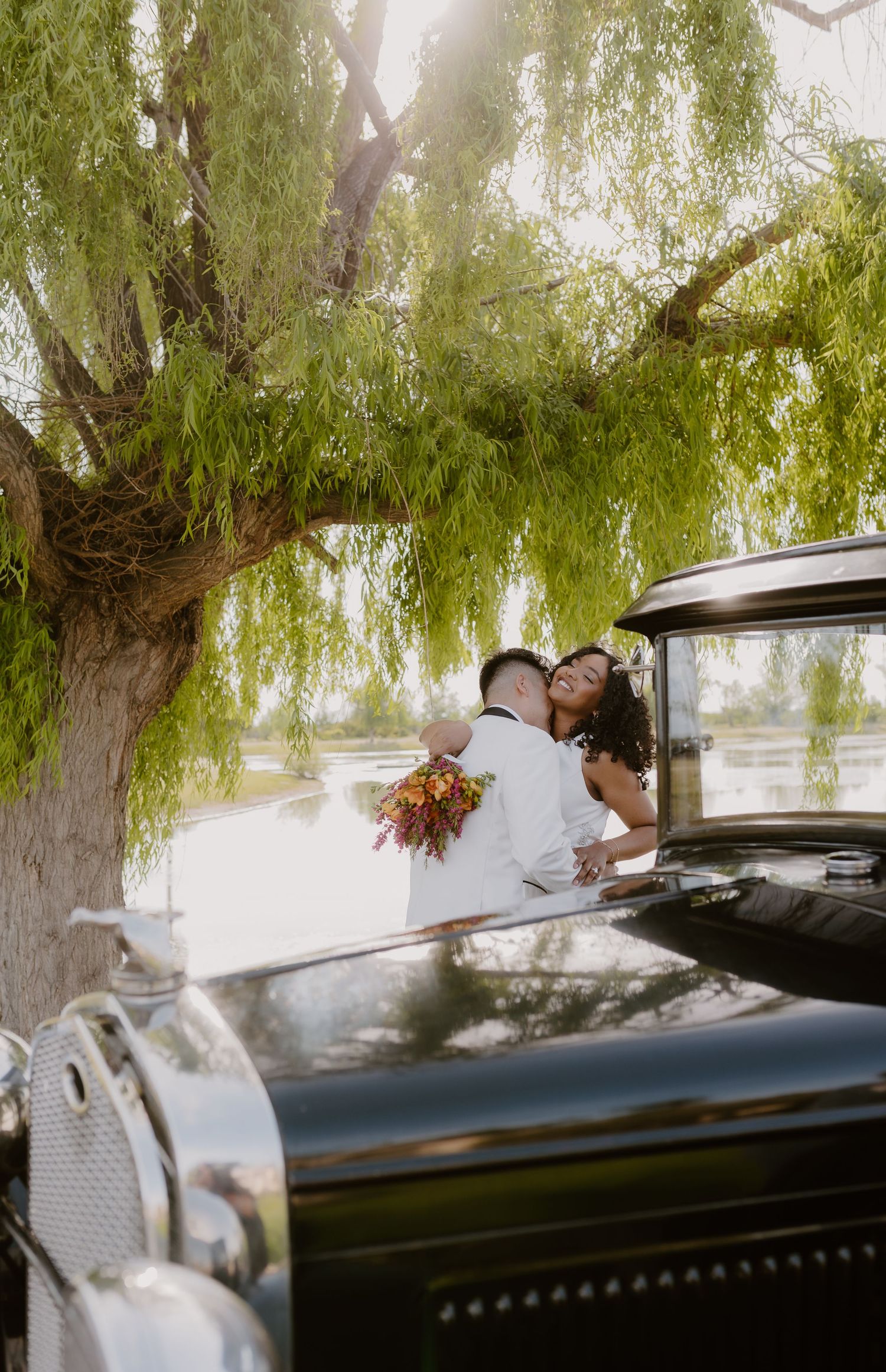Bride and groom embrace under a weeping willow tree beside a vintage black classic car near a serene lake.