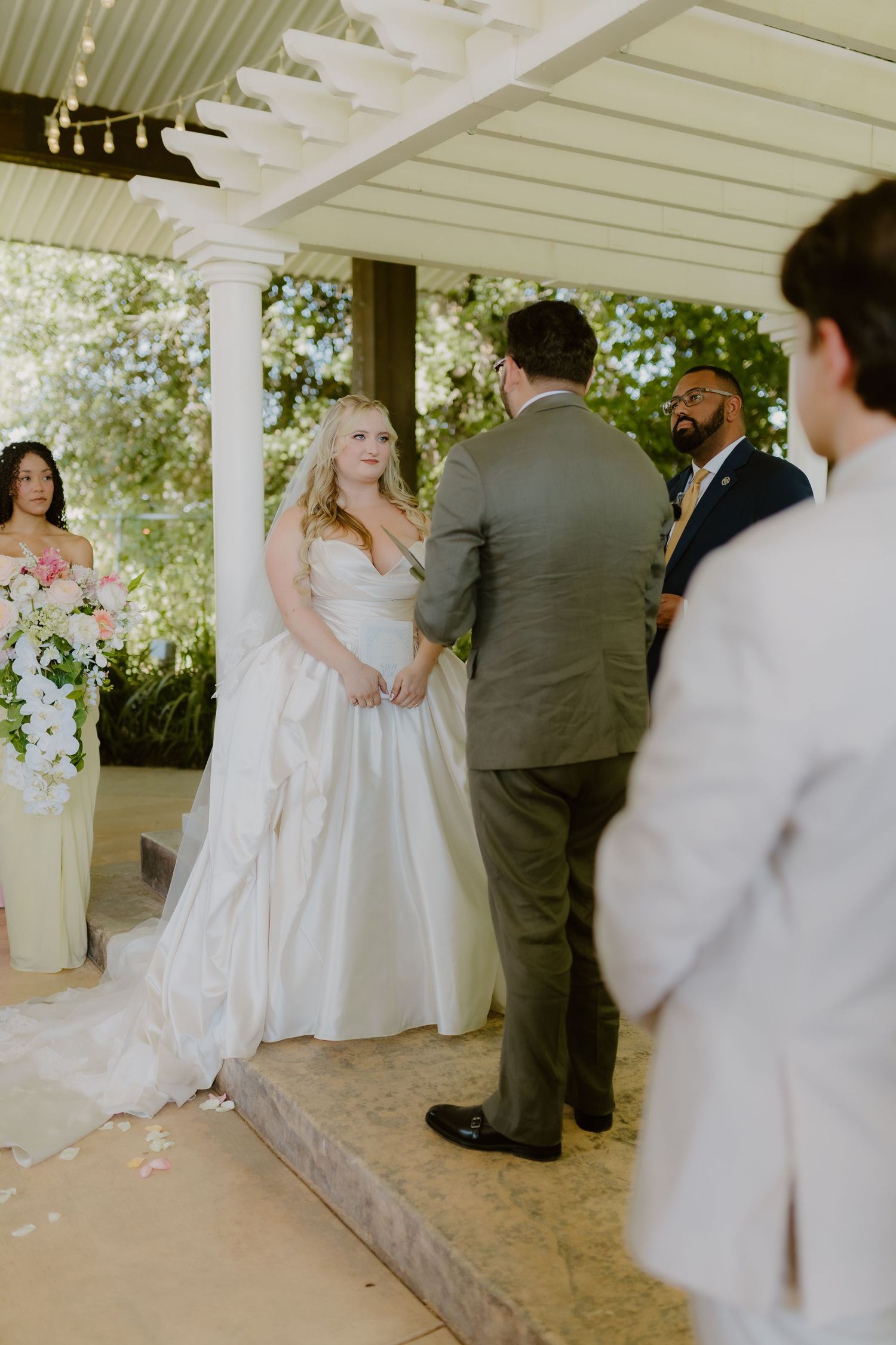 Blonde bride in white gown holding hands with groom at outdoor pergola wedding ceremony.