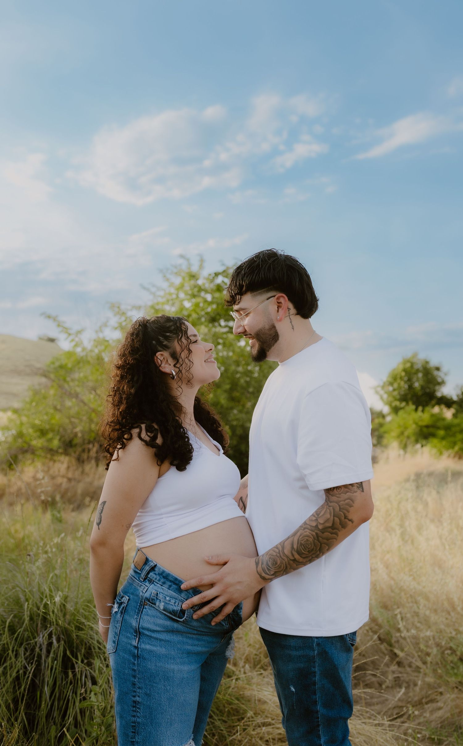 A smiling couple cradles a pregnant belly together while standing in a sunny golden grass field.