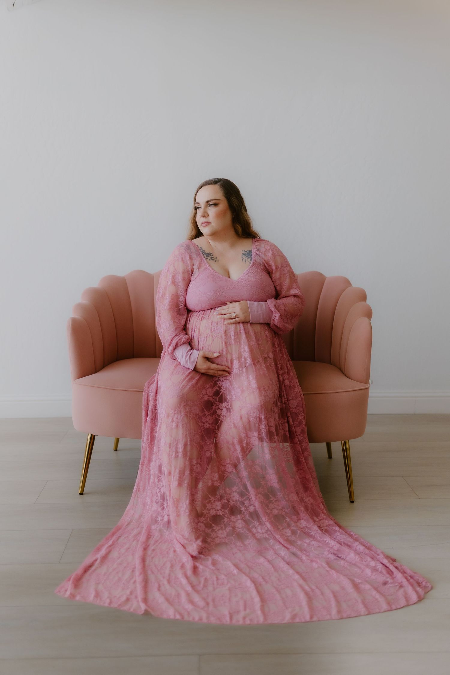 Pregnant woman in elegant pink floral maxi gown seated before a blush velvet chair in studio.