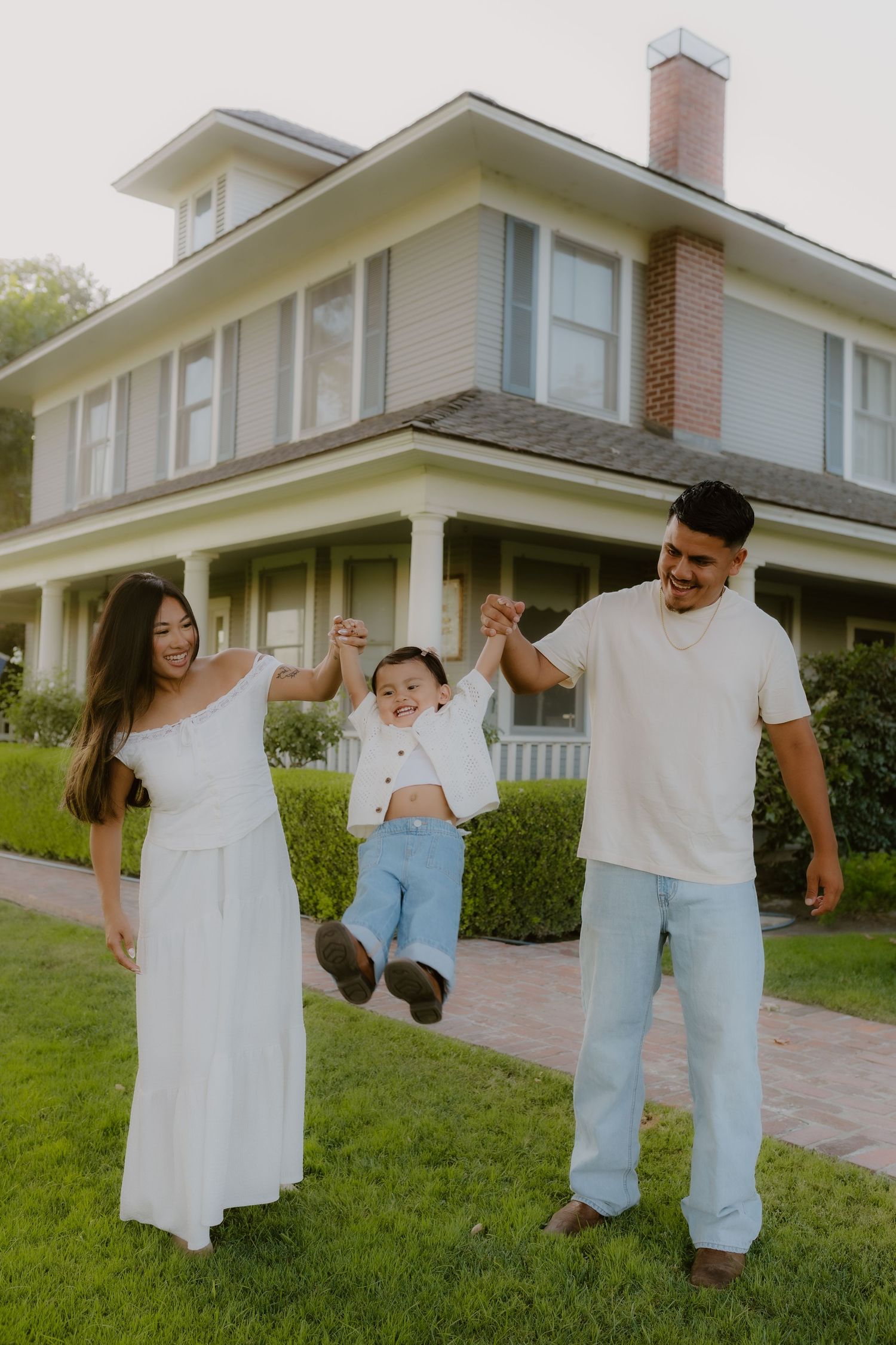 Young family joyfully swinging toddler between parents on lawn outside classic two-story home. California, The Charm House