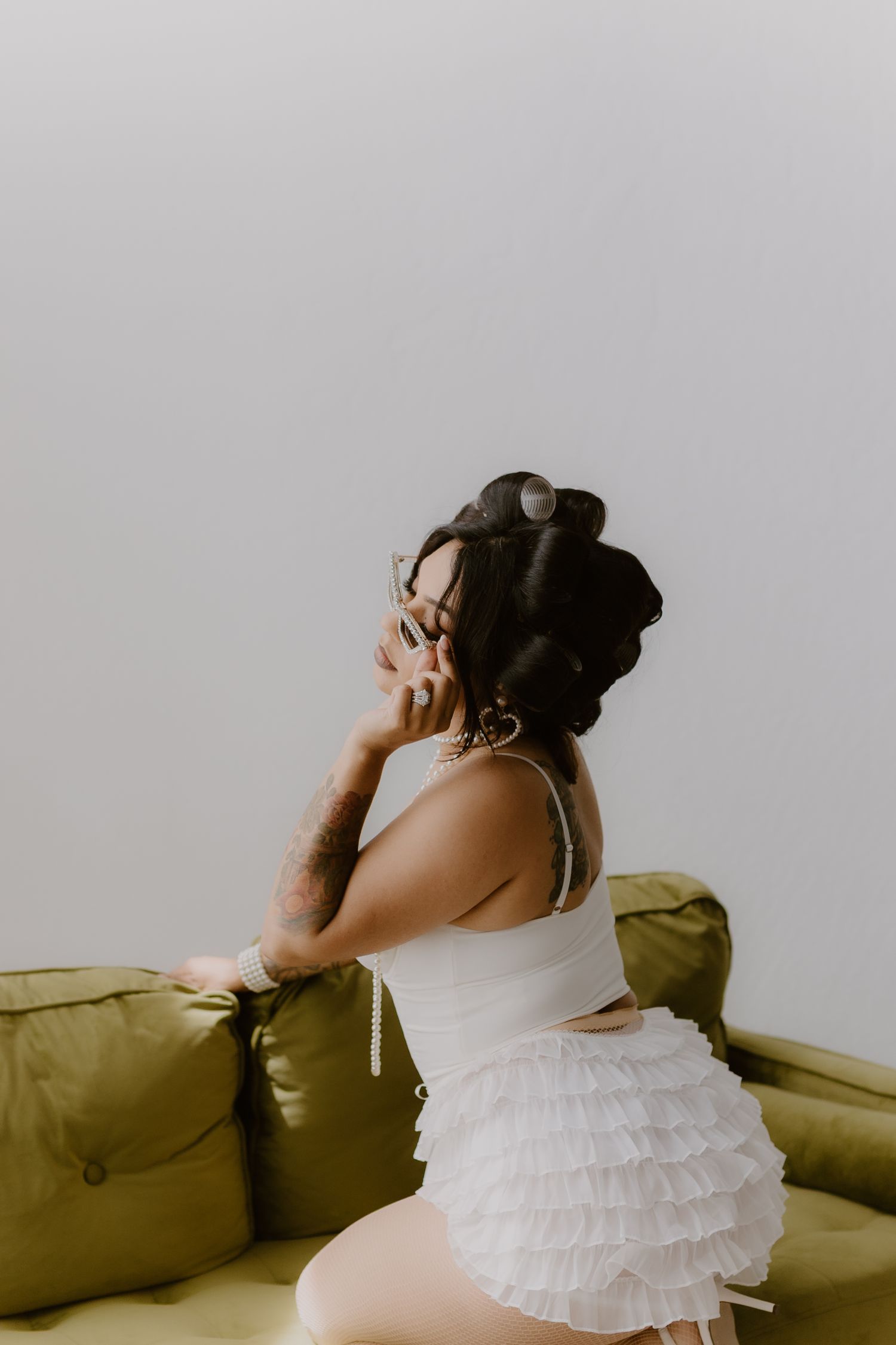 Woman in white ruffled outfit with hair rollers poses on green velvet sofa in bright room. Tulare, California