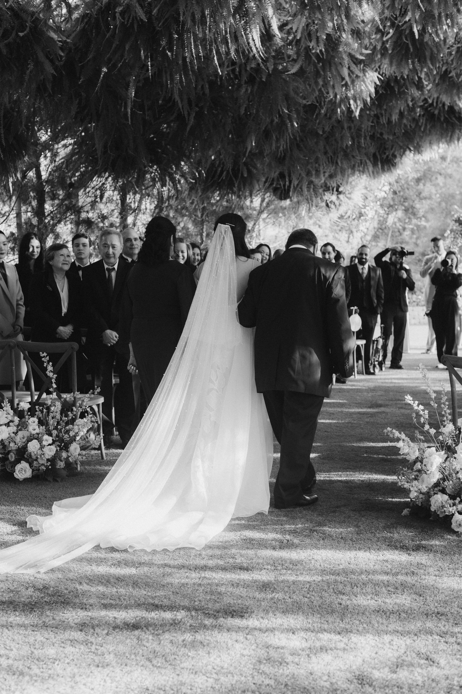 Bride walking down outdoor wedding aisle in flowing veil with escort, guests watching among trees. Ethereal Gardens, California, Southern