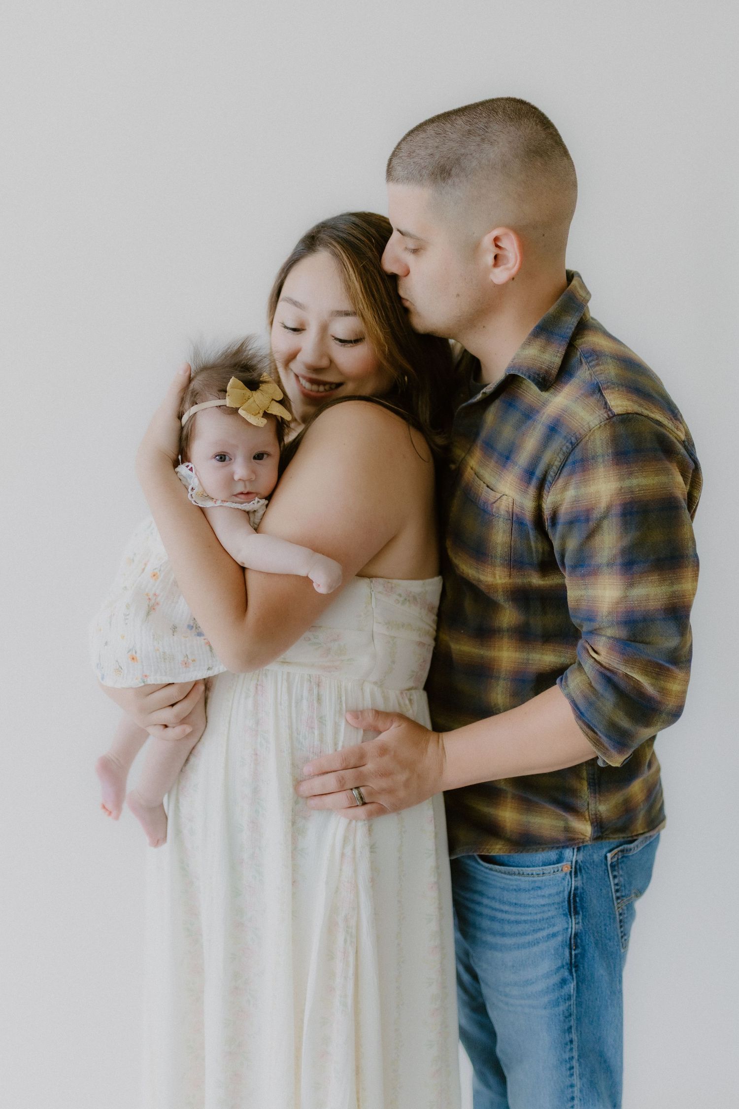A tender family portrait of a mother holding her baby girl as father kisses her head from behind.
