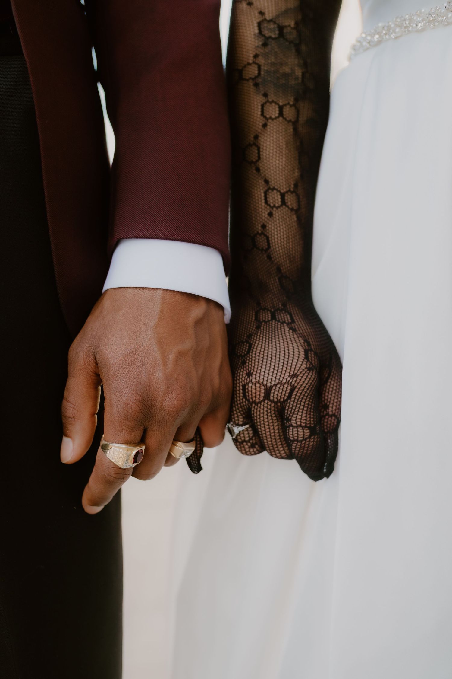 Close-up of interracial wedding couple holding hands, bride wearing black Gucci lace gloves and groom in burgundy suit.