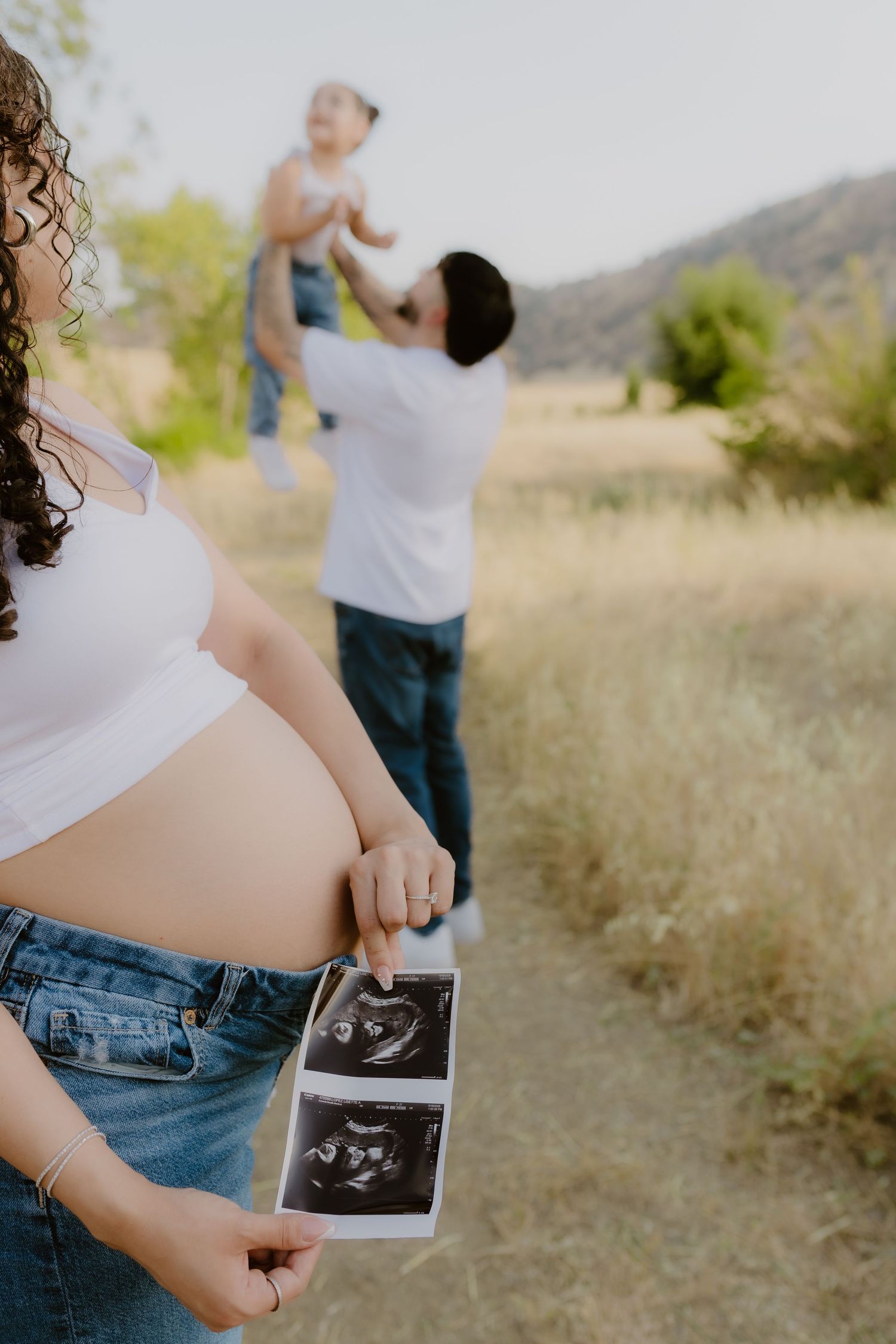 Pregnant woman holds ultrasound photos while a man lifts a child in a sunny outdoor field behind her.
