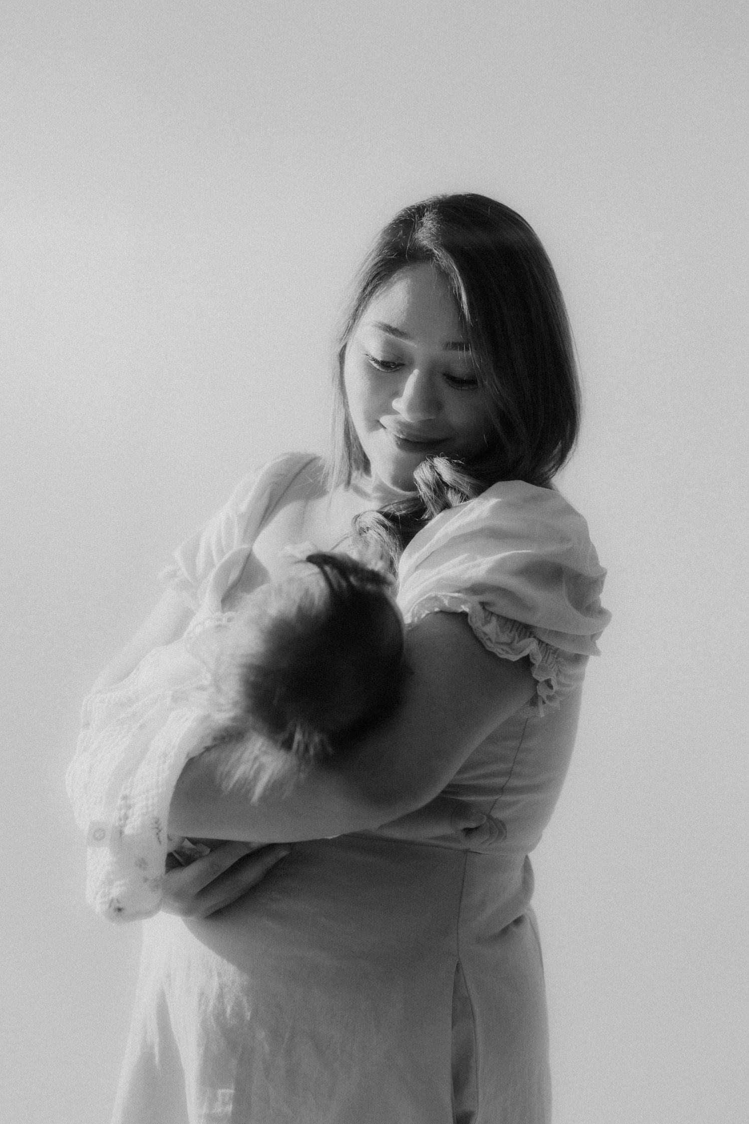 Tender black and white portrait of mother in white dress standing and cradling sleeping newborn baby.