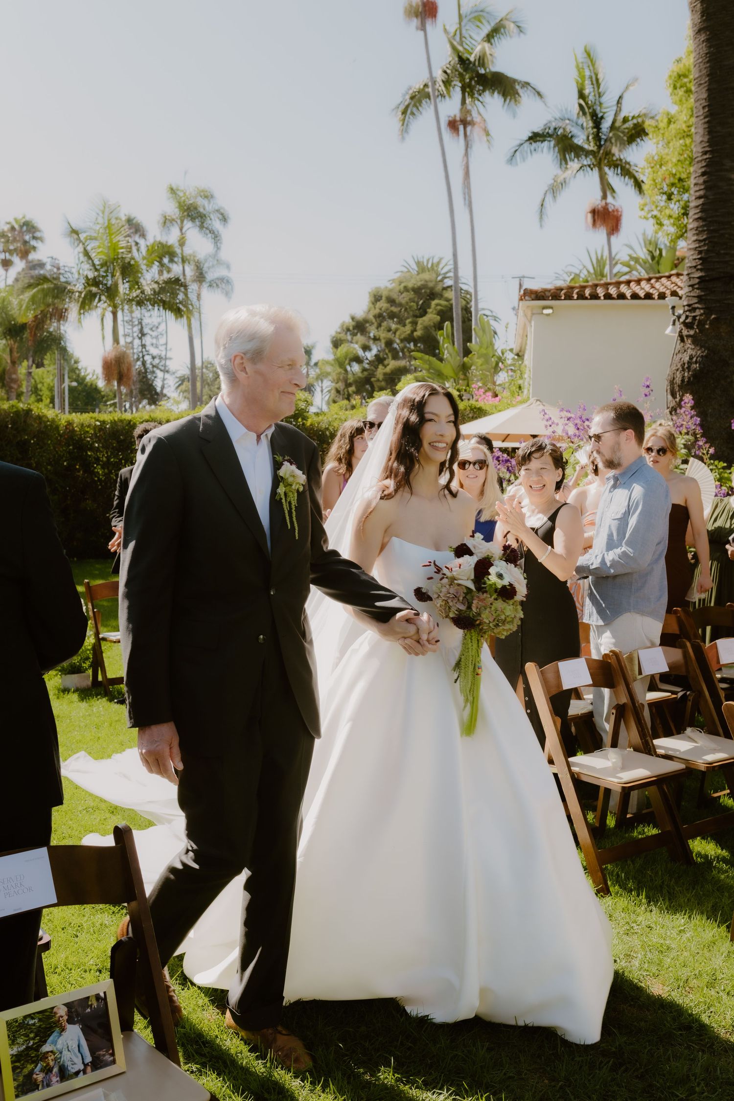 Bride in white gown walks down outdoor aisle with older gentleman escort at sunny garden wedding.