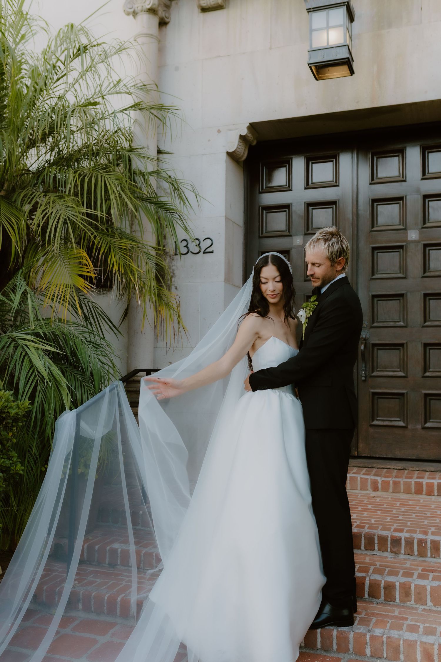 Groom tenderly holds bride as her long veil cascades beside them on brick steps of an elegant building.