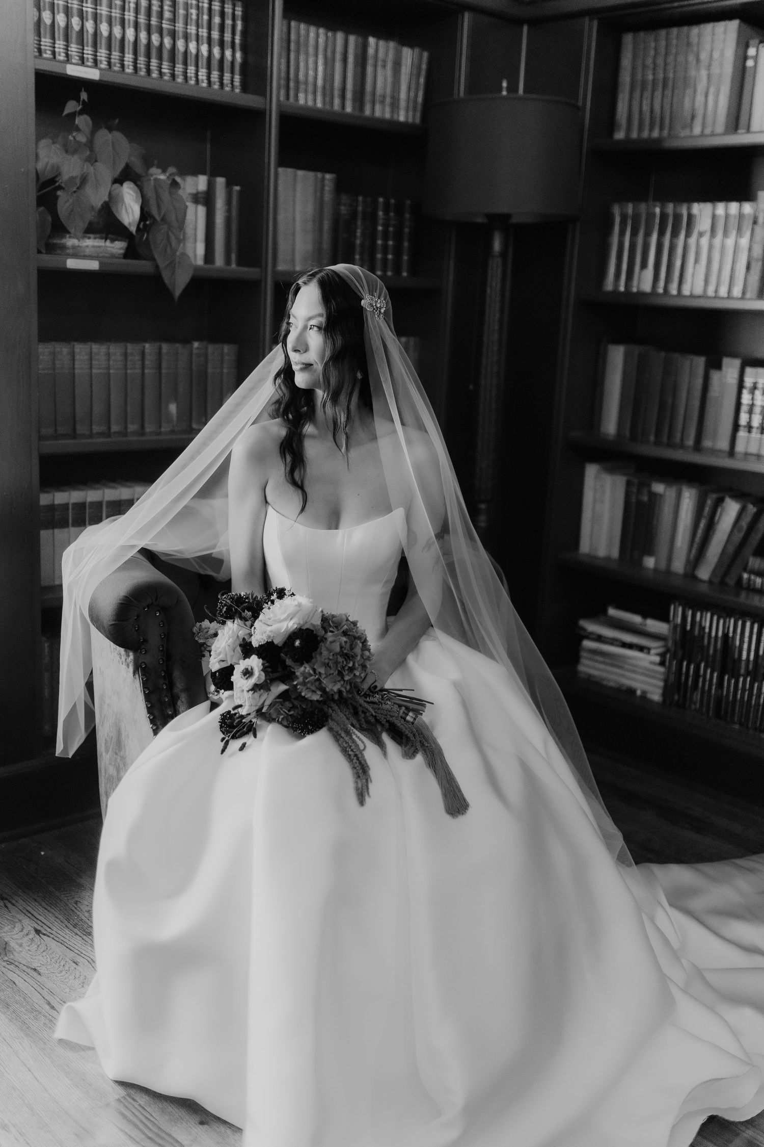 Black and white portrait of bride in ball gown holding bouquet while seated in library chair.