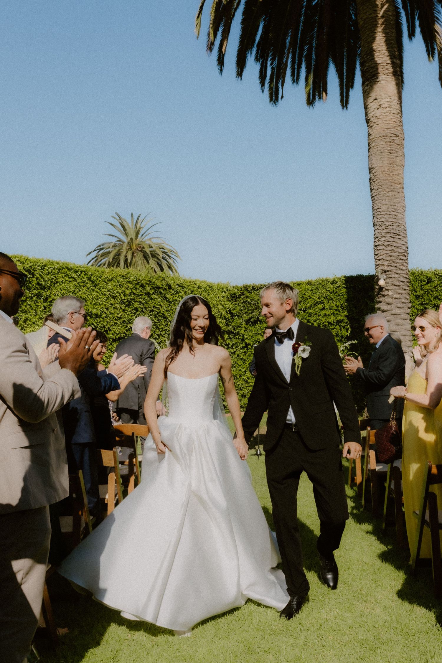 Newlyweds walk joyfully down outdoor aisle lined with applauding guests beneath a tall palm tree. California, Santa Barbara wedding