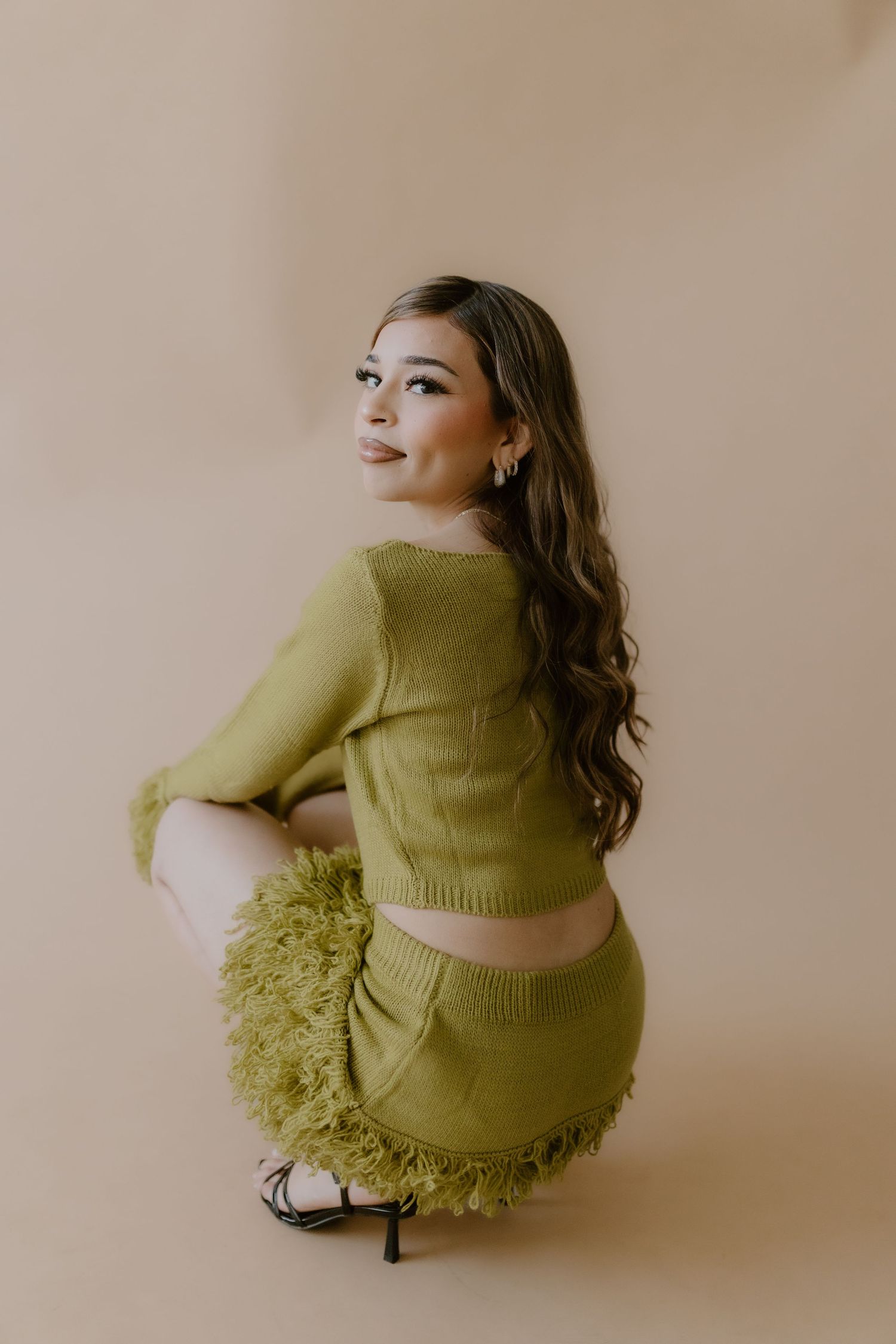 Woman in olive green feathered knit set sits on heels looking over shoulder against warm beige background.