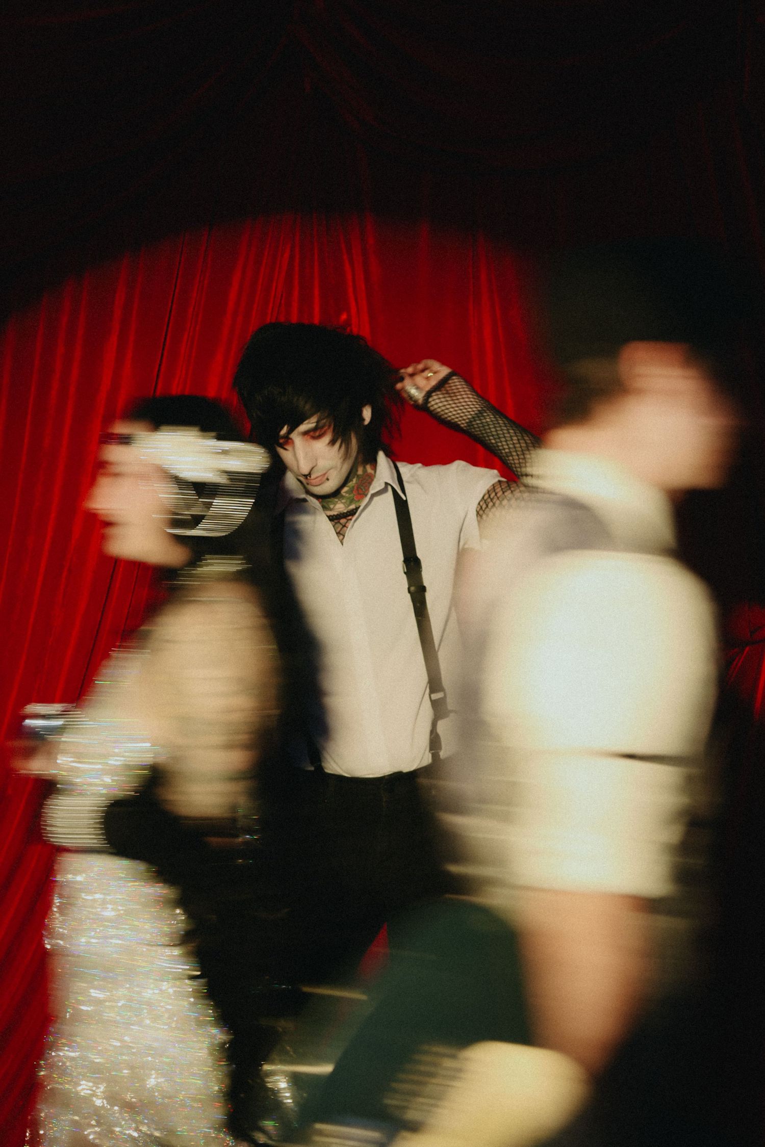 Motion-blurred artistic shot of goth guitarist performing before red stage curtain.