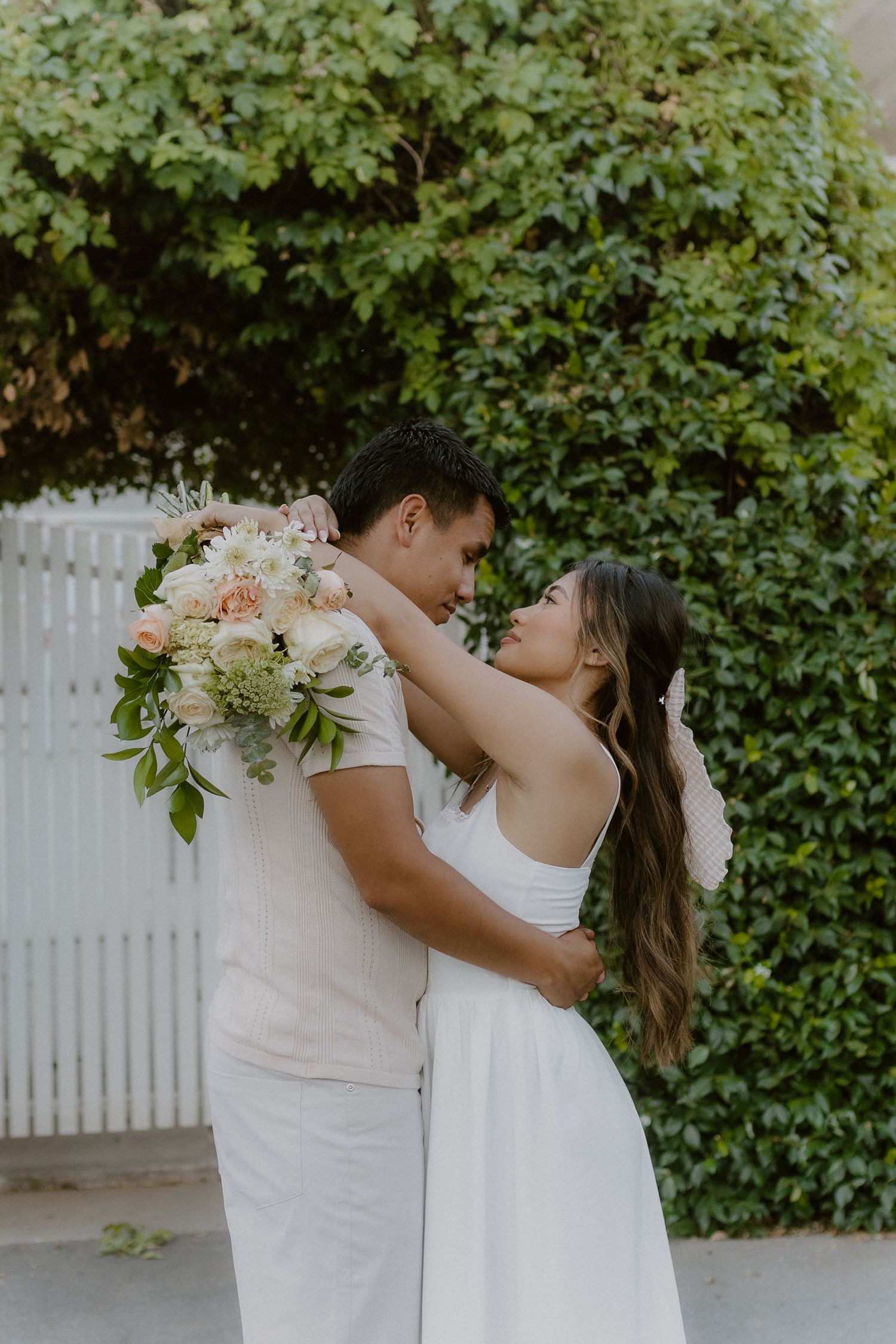 Bride and groom sharing a tender moment with a beautiful white and peach bouquet outdoors.