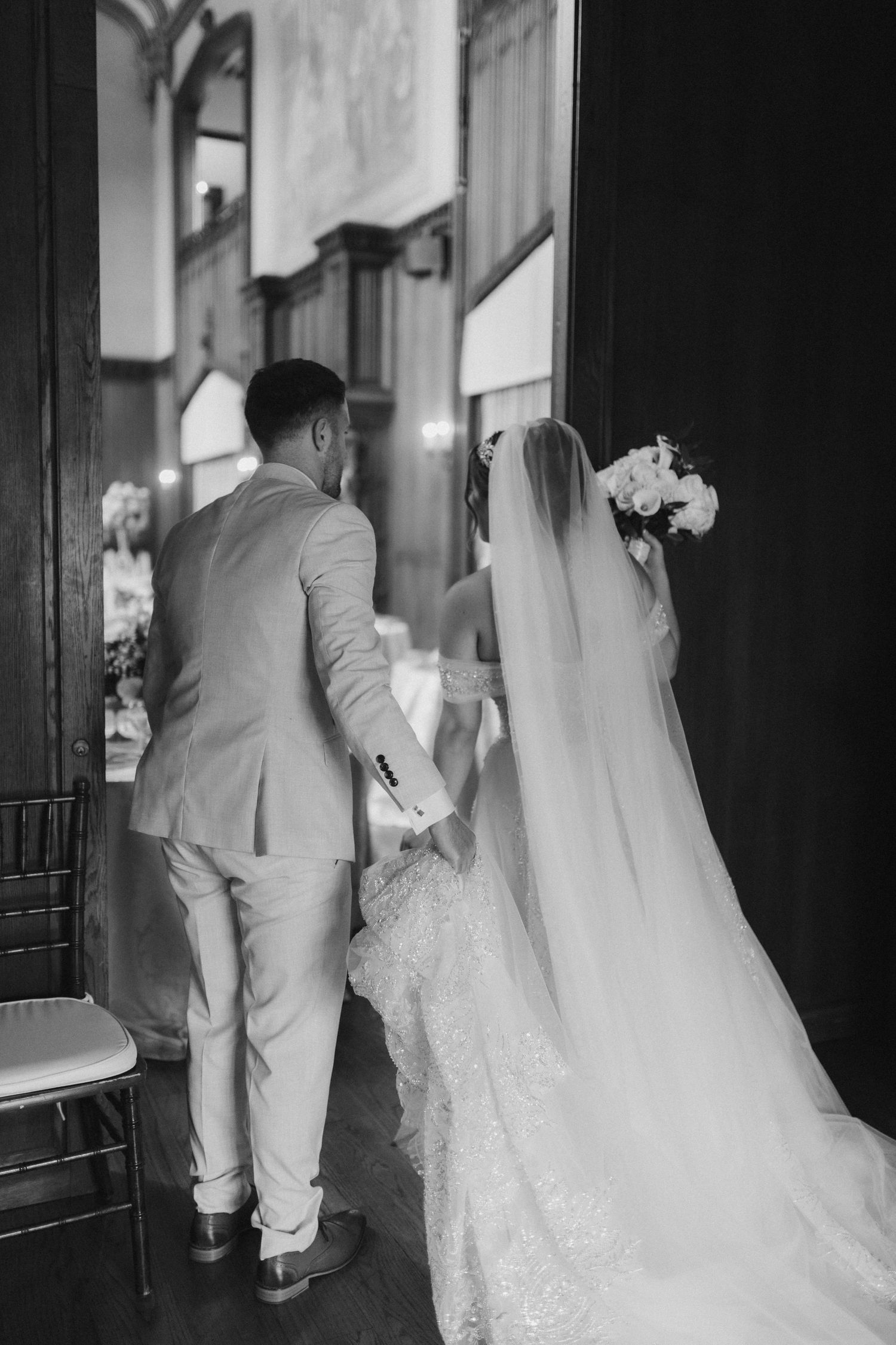 Black and white photo of bride and groom holding hands walking into ceremony through ornate wooden doors.