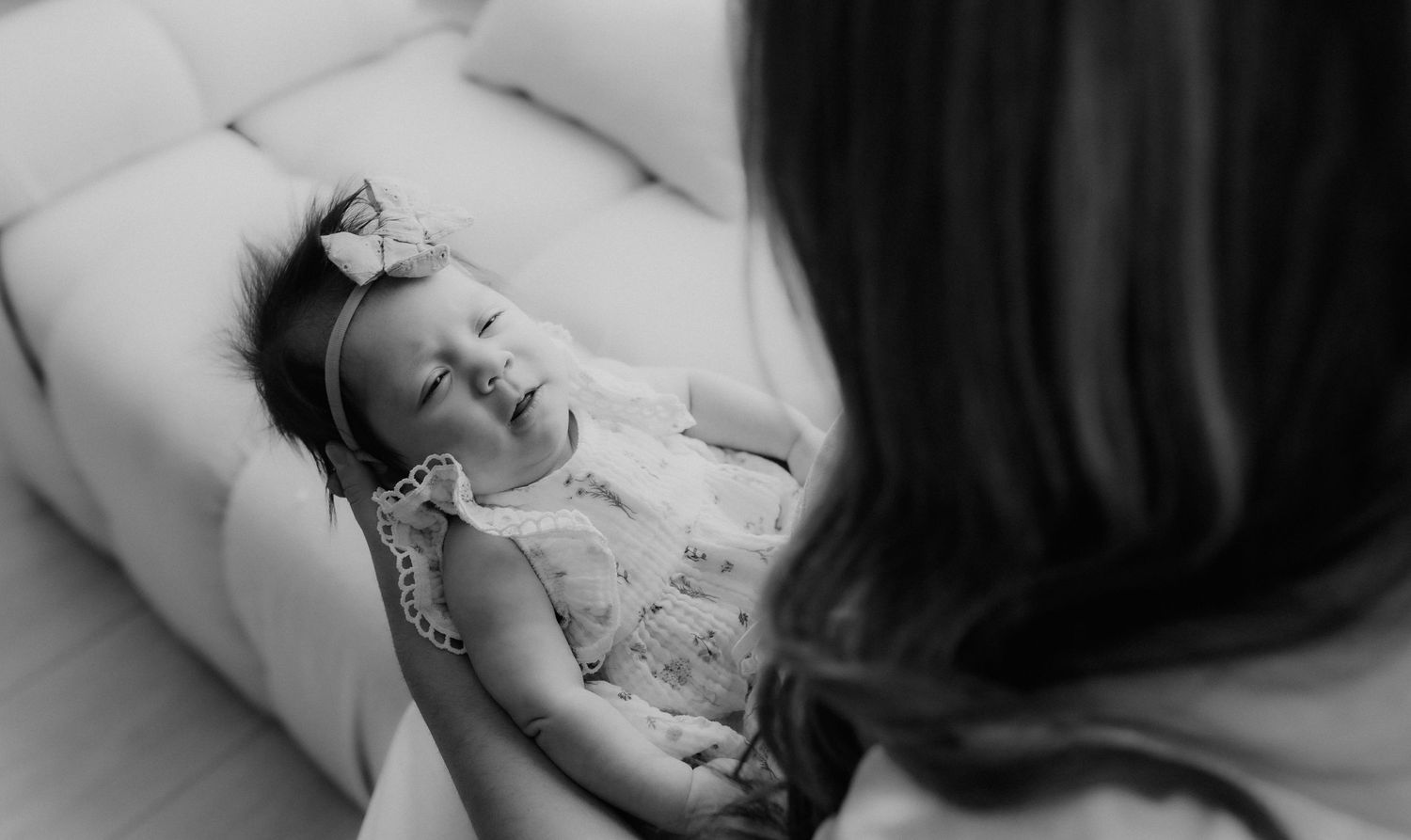 Black and white image of a baby girl in a bow headband smiling up at her mother on a cream sofa.