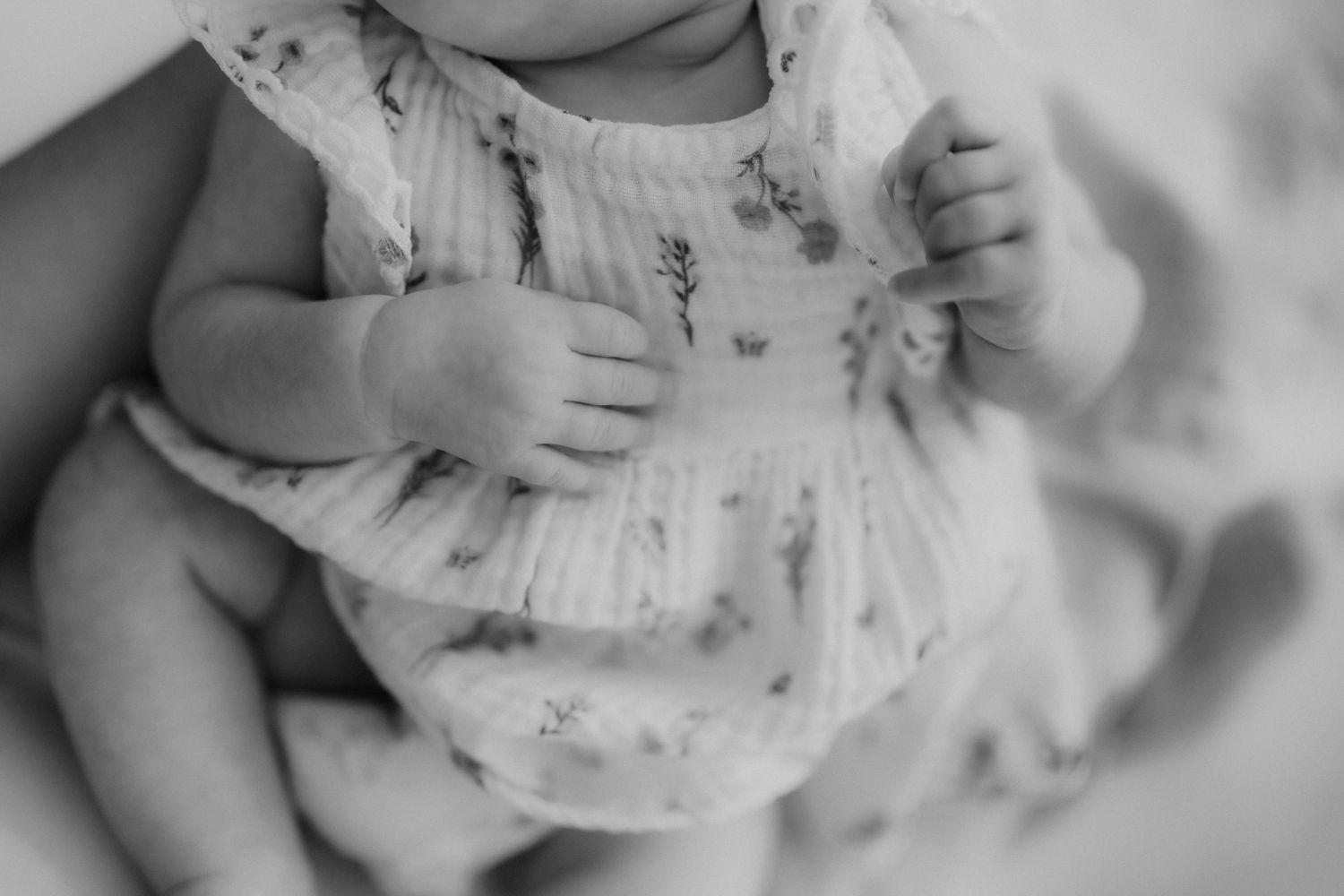 Black and white close-up of a newborn baby in a floral romper with tiny folded hands.