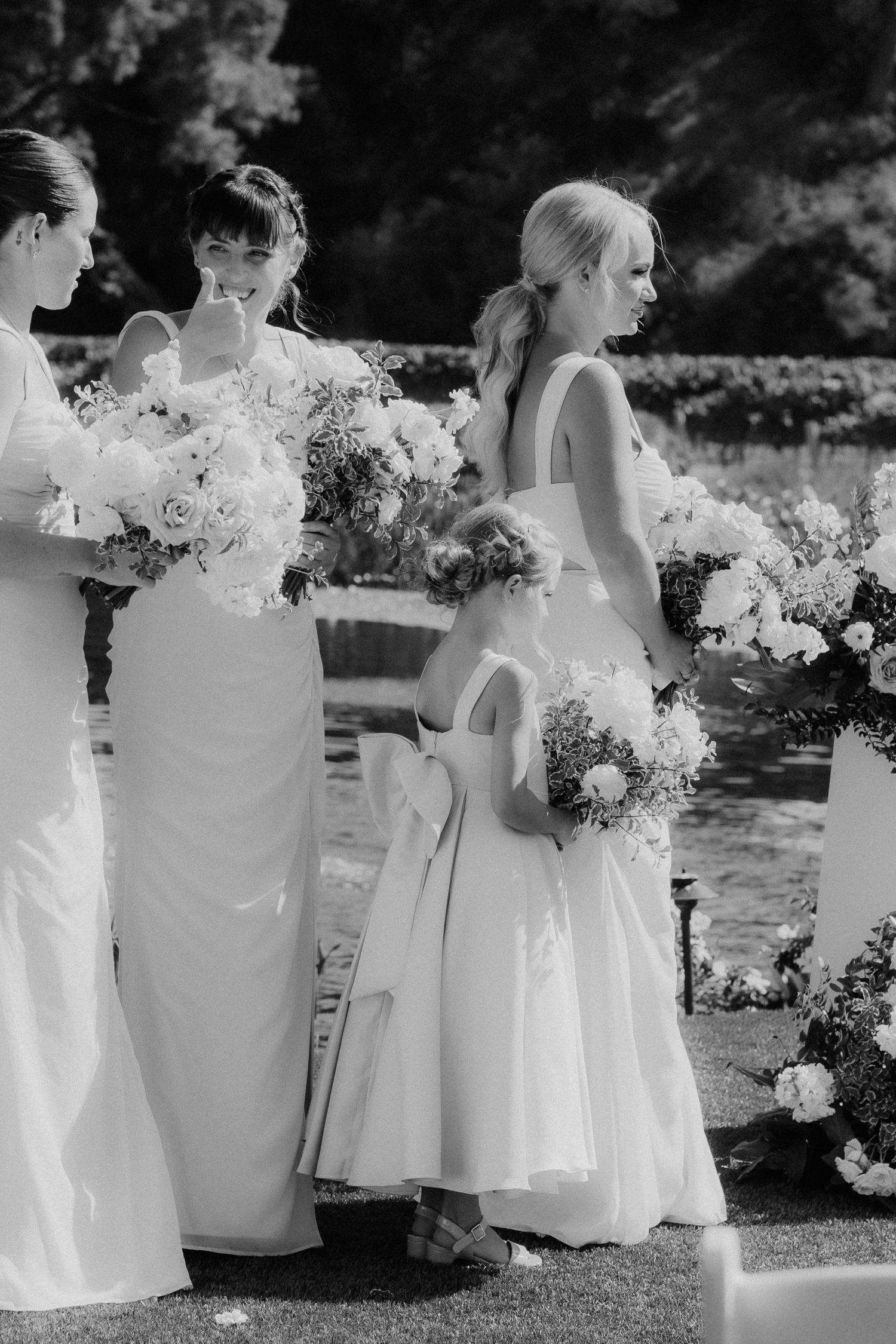 Black and white photo of bridesmaids and flower girl holding bouquets at an outdoor wedding ceremony.