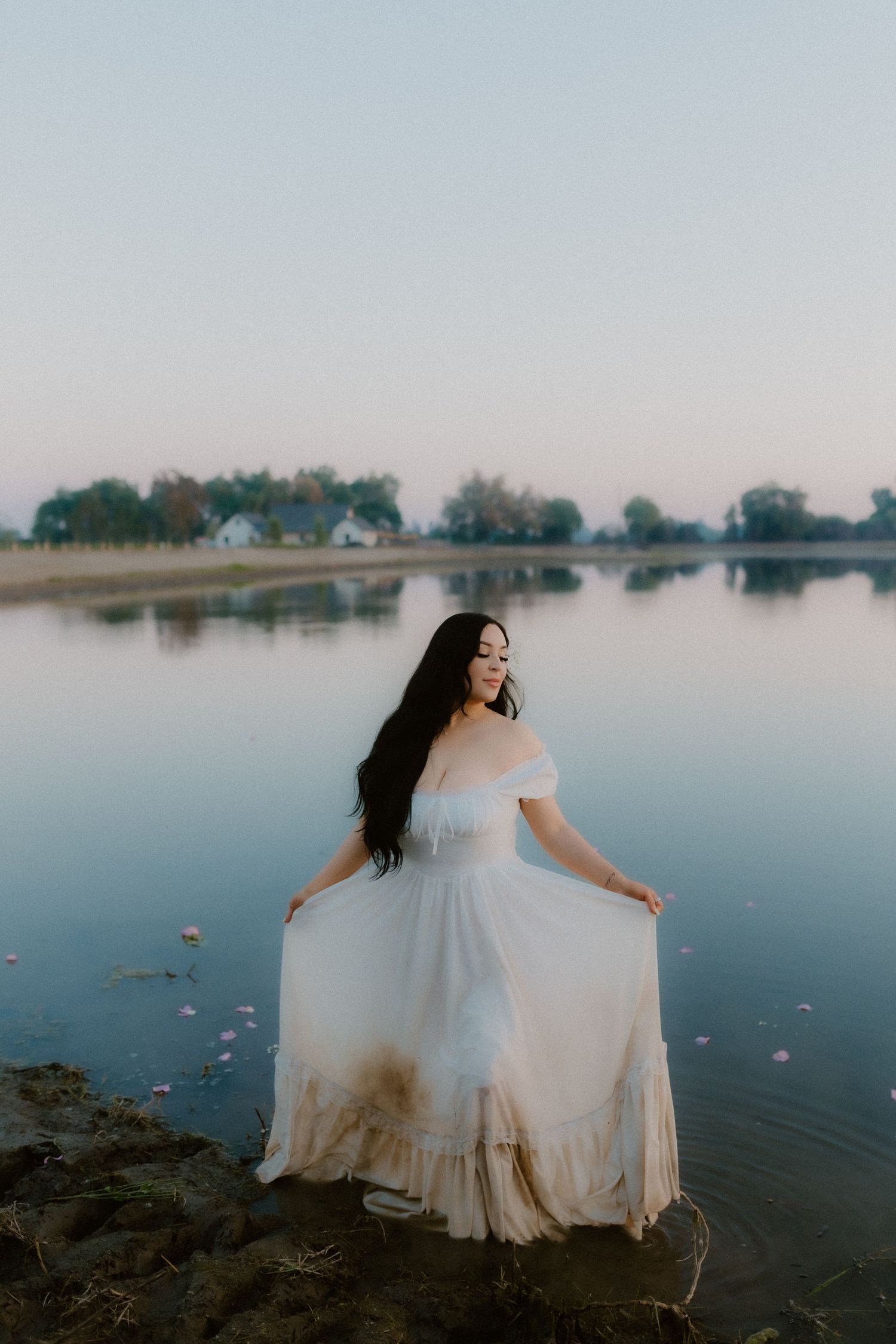 Bride in off-shoulder white gown holding skirt while standing at edge of tranquil lake.