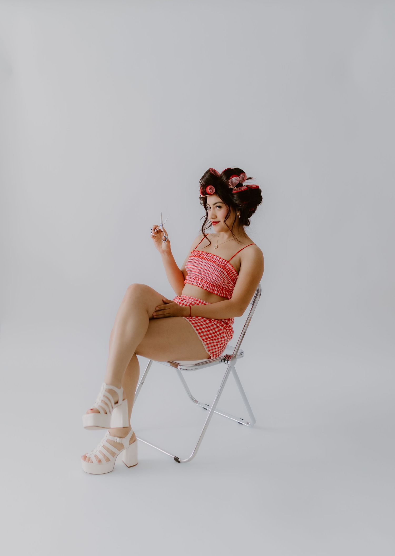 Woman in red gingham two-piece set with hair rollers sits on folding chair in white studio.