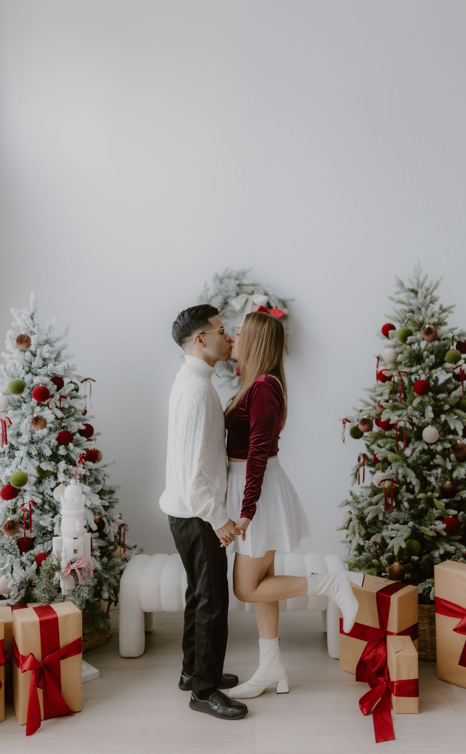 Couple shares a kiss surrounded by decorated Christmas trees and wrapped gifts in a bright white studio.