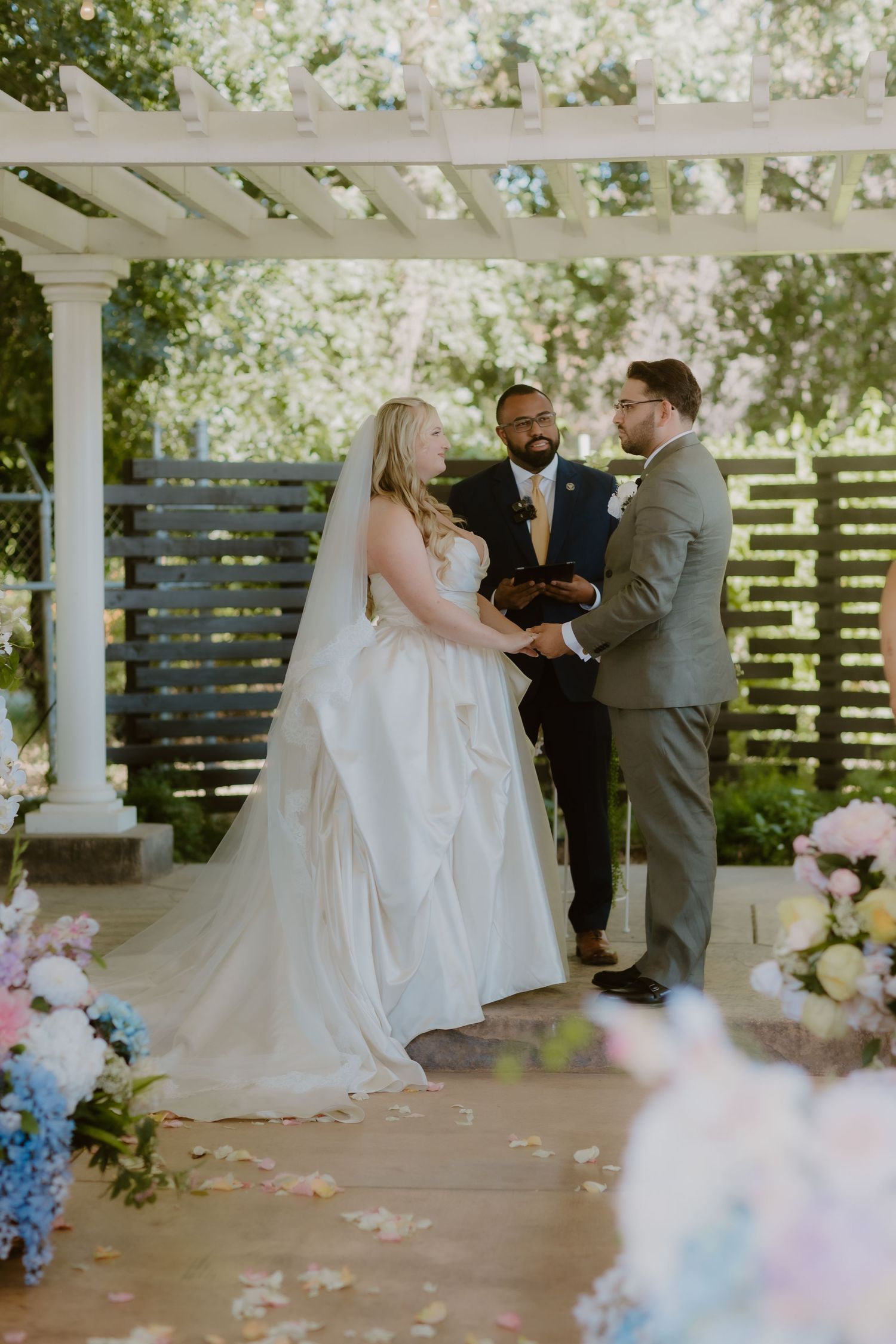 Bride and groom exchange vows under a white pergola decorated with pink and blue flowers at an outdoor ceremony.
