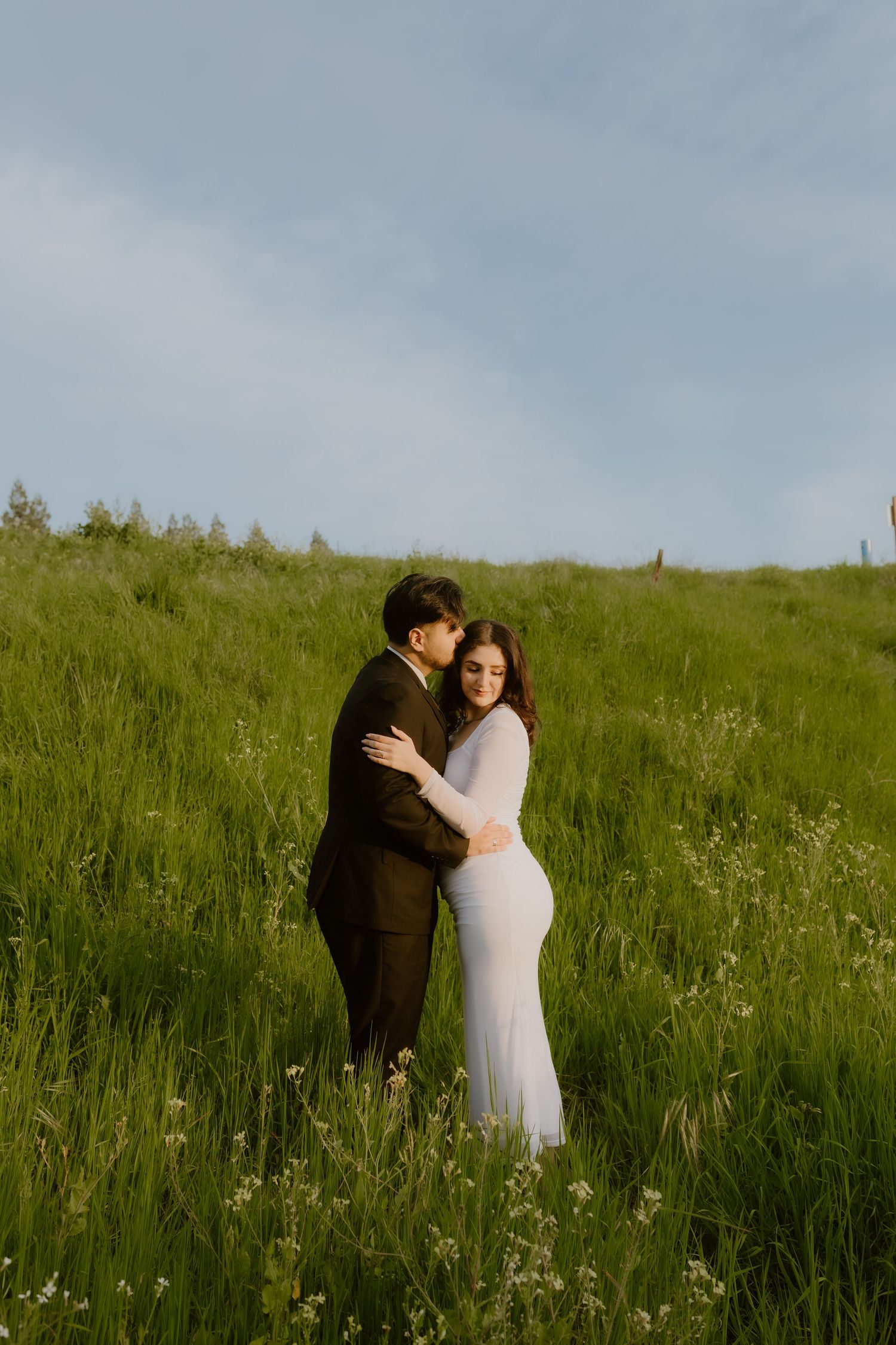 Couple in formal wear embracing in a lush green meadow under a blue sky.